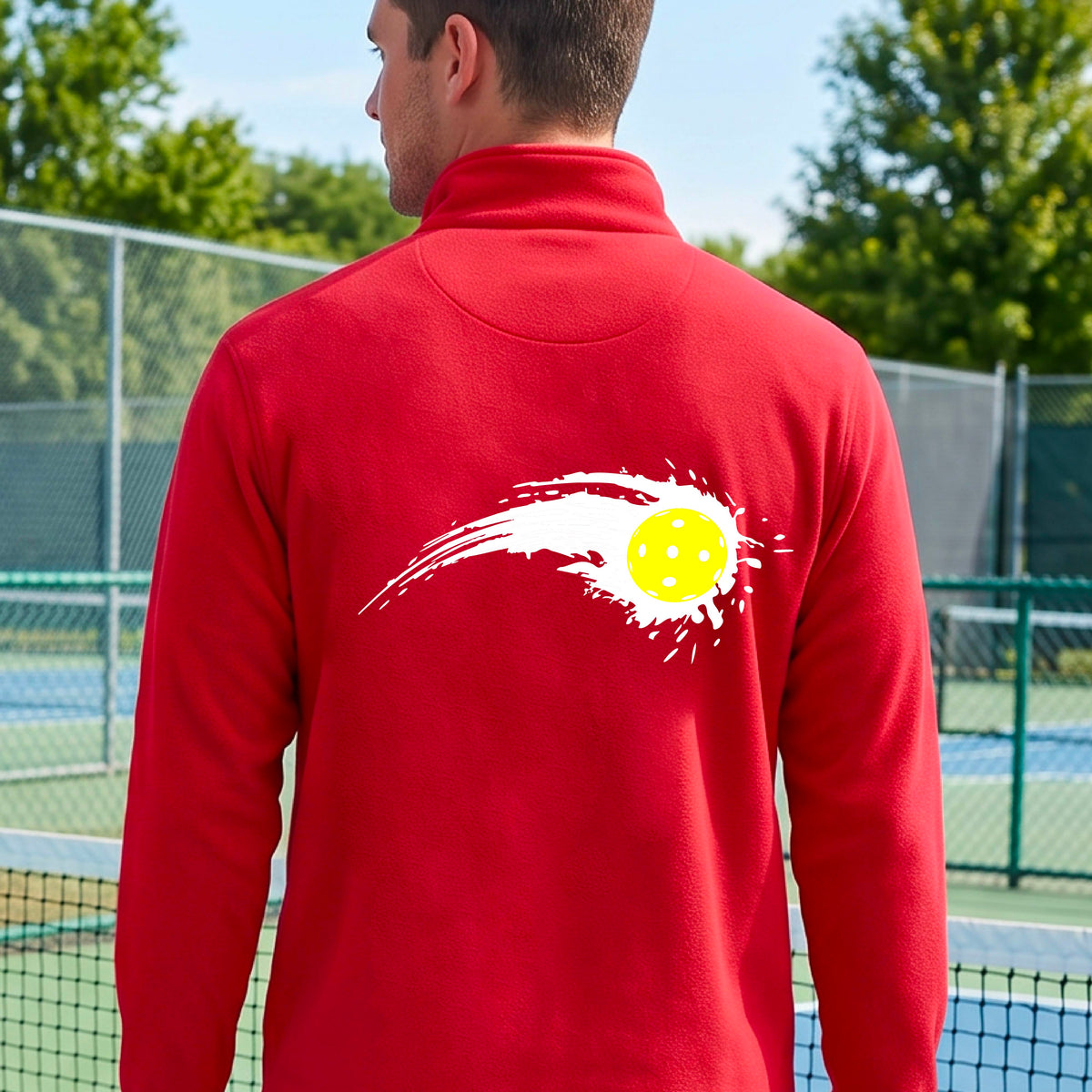 A man wearing a red long-sleeve quarter-zip shirt with a white pickleball graphic of a yellow pickleball with a motion trail effect, displayed in front of a pickleball court. 