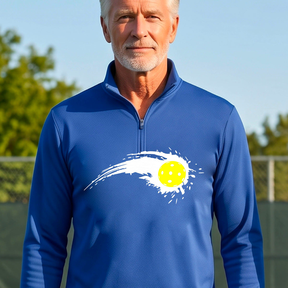A man wearing a royal blue long-sleeve quarter-zip shirt with a white pickleball graphic of a yellow pickleball with a motion trail effect, displayed in front of a pickleball court. 