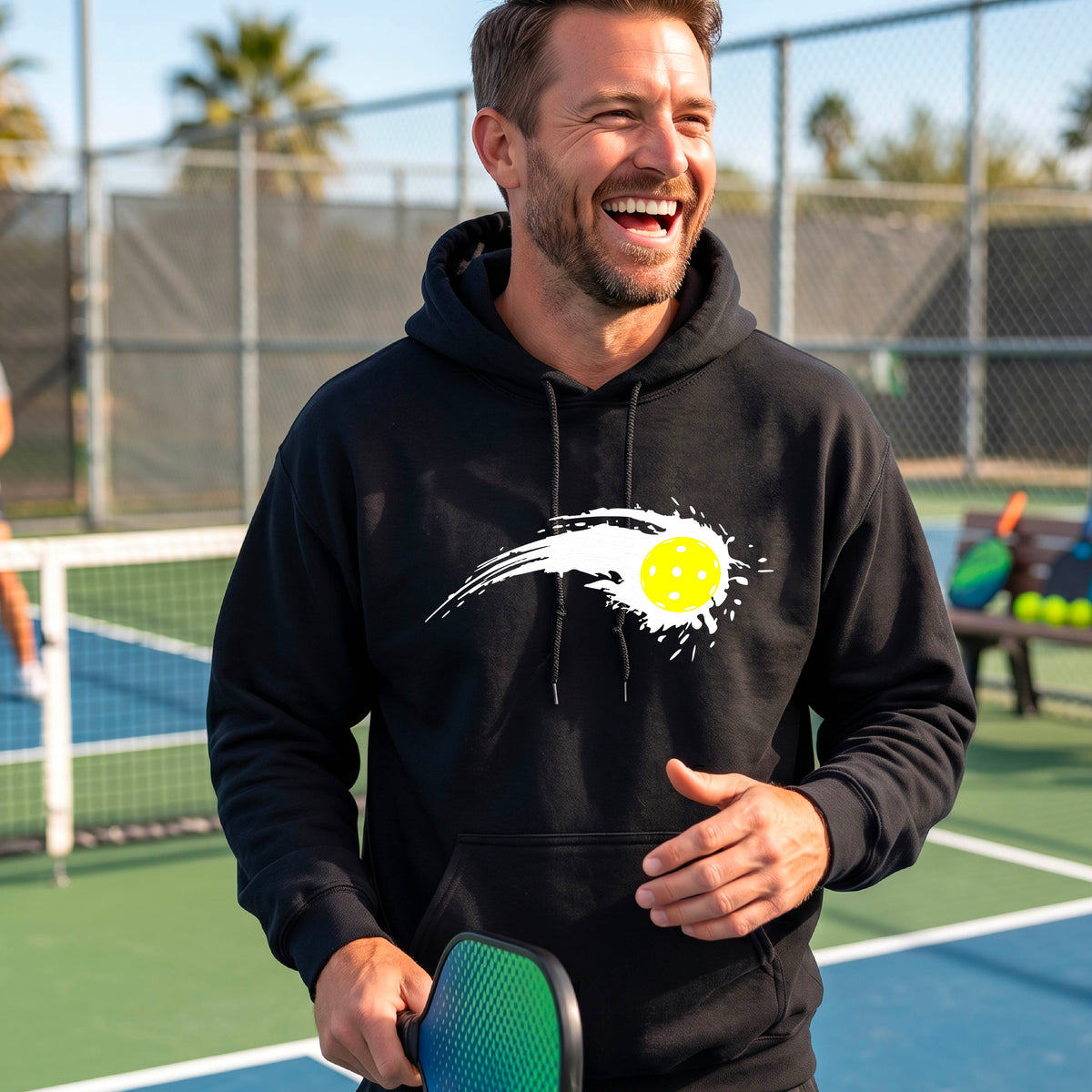 A man wearing a black unisex hoodie pullover with a white pickleball graphic of a yellow pickleball with a motion trail effect, displayed in front of a pickleball court. 