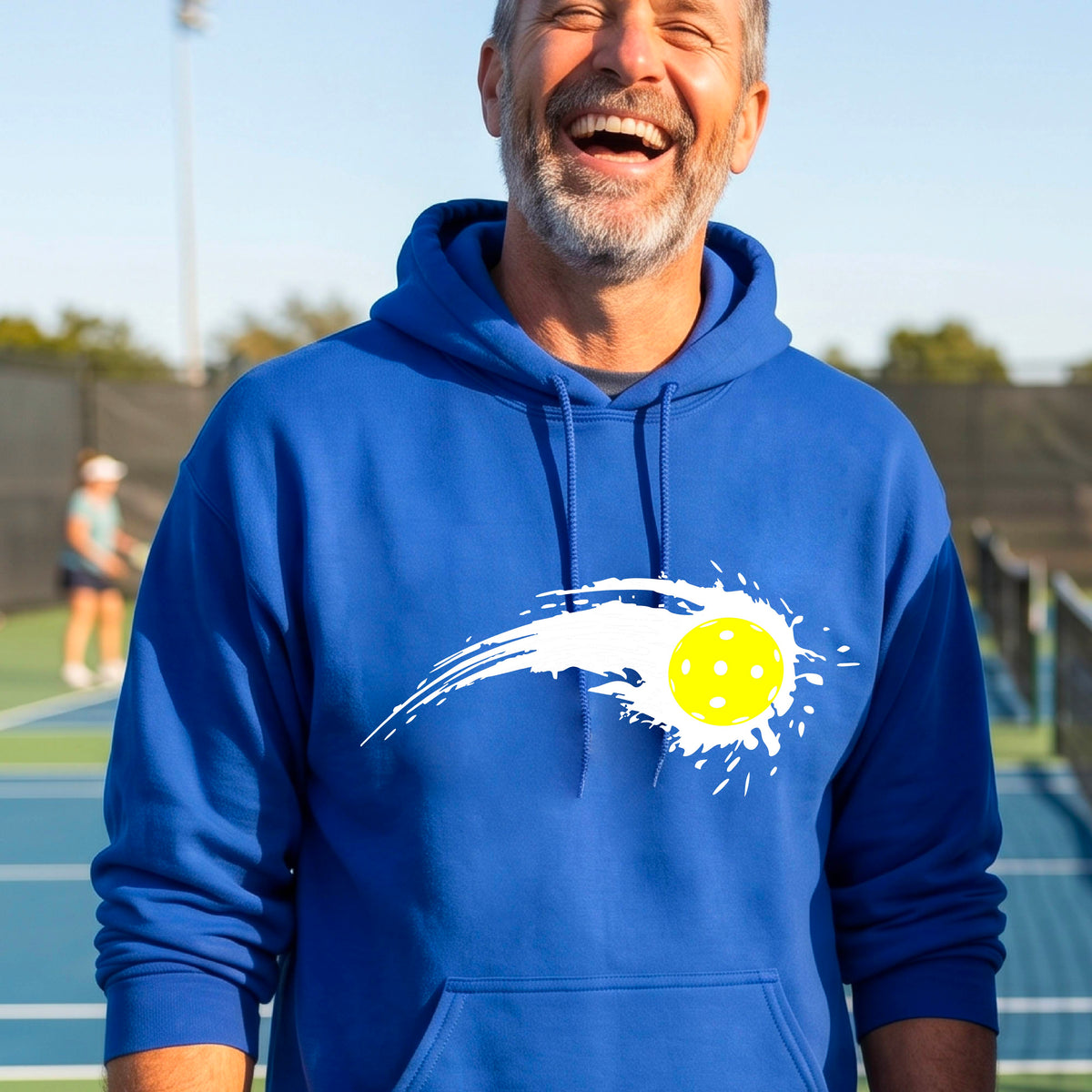 A man wearing a royal blue unisex hoodie pullover with a white pickleball graphic of a yellow pickleball with a motion trail effect, displayed in front of a pickleball court. 