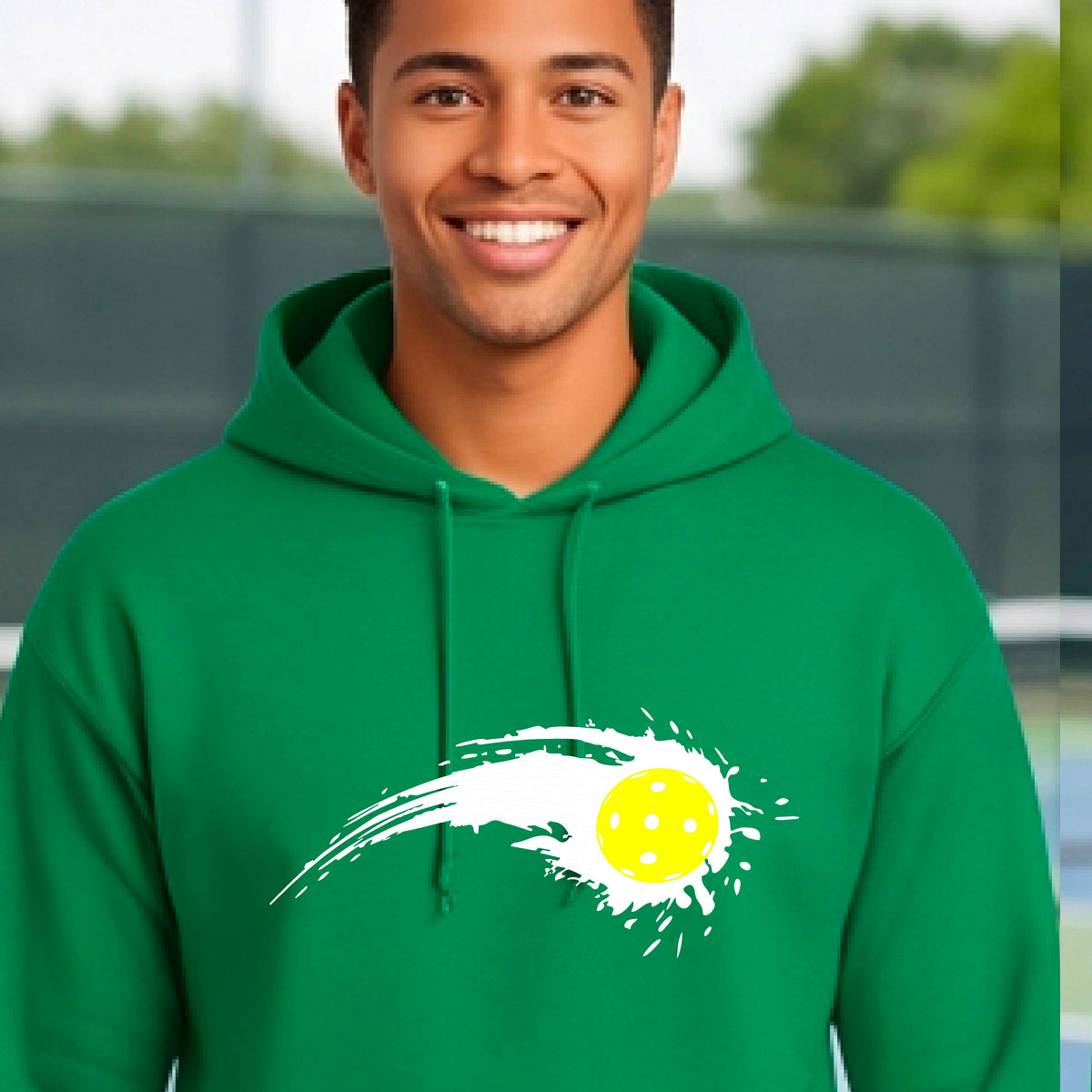 A man wearing a green unisex hoodie pullover with a white pickleball graphic of a yellow pickleball with a motion trail effect, displayed in front of a pickleball court. 