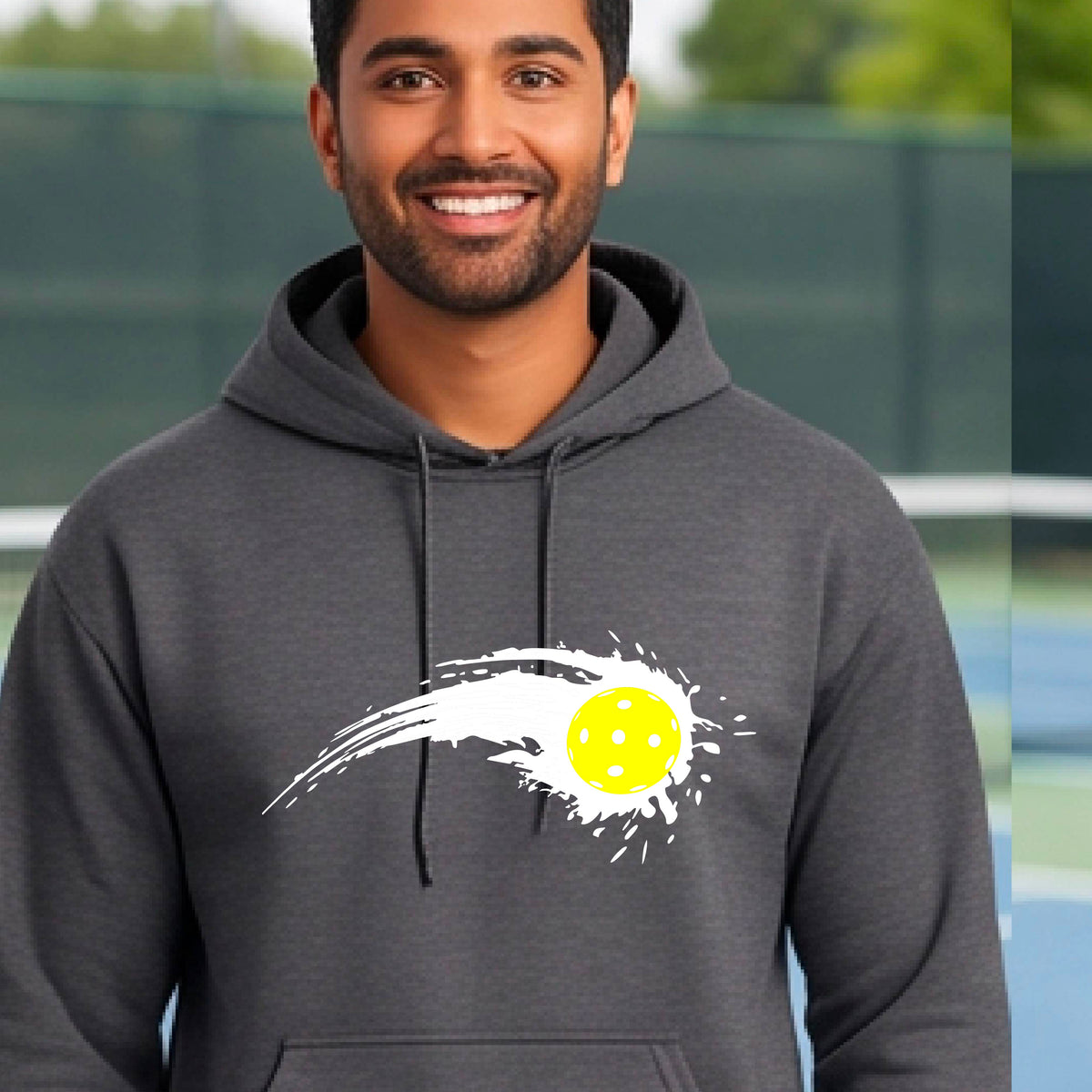 A man wearing a dark gray unisex hoodie pullover with a white pickleball graphic of a yellow pickleball with a motion trail effect, displayed in front of a pickleball court. 