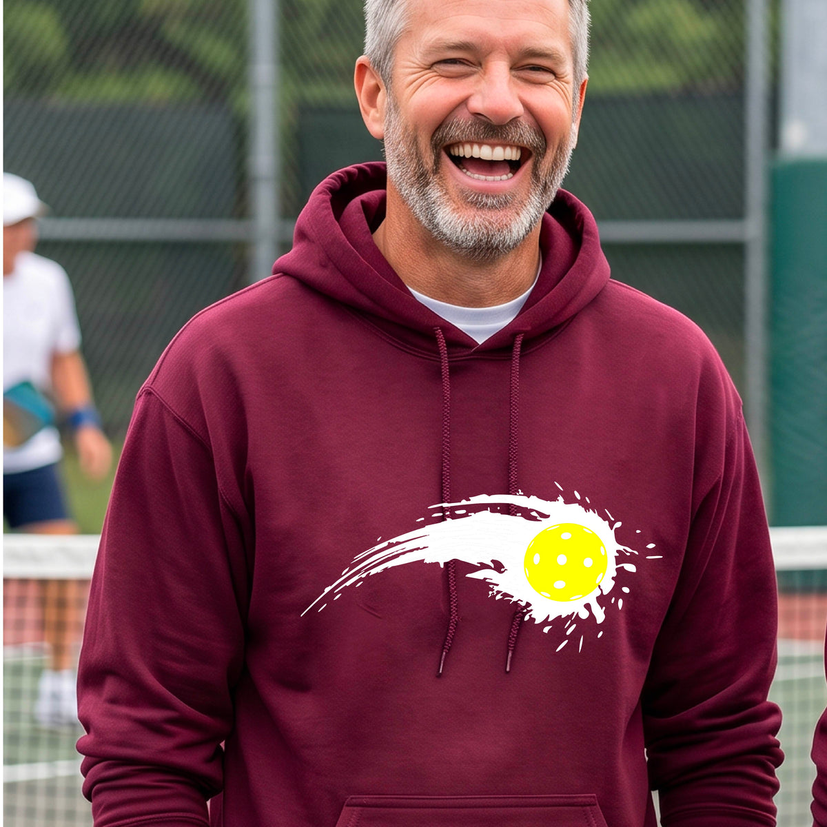 A man wearing a maroon unisex hoodie pullover with a white pickleball graphic of a yellow pickleball with a motion trail effect, displayed in front of a pickleball court. 
