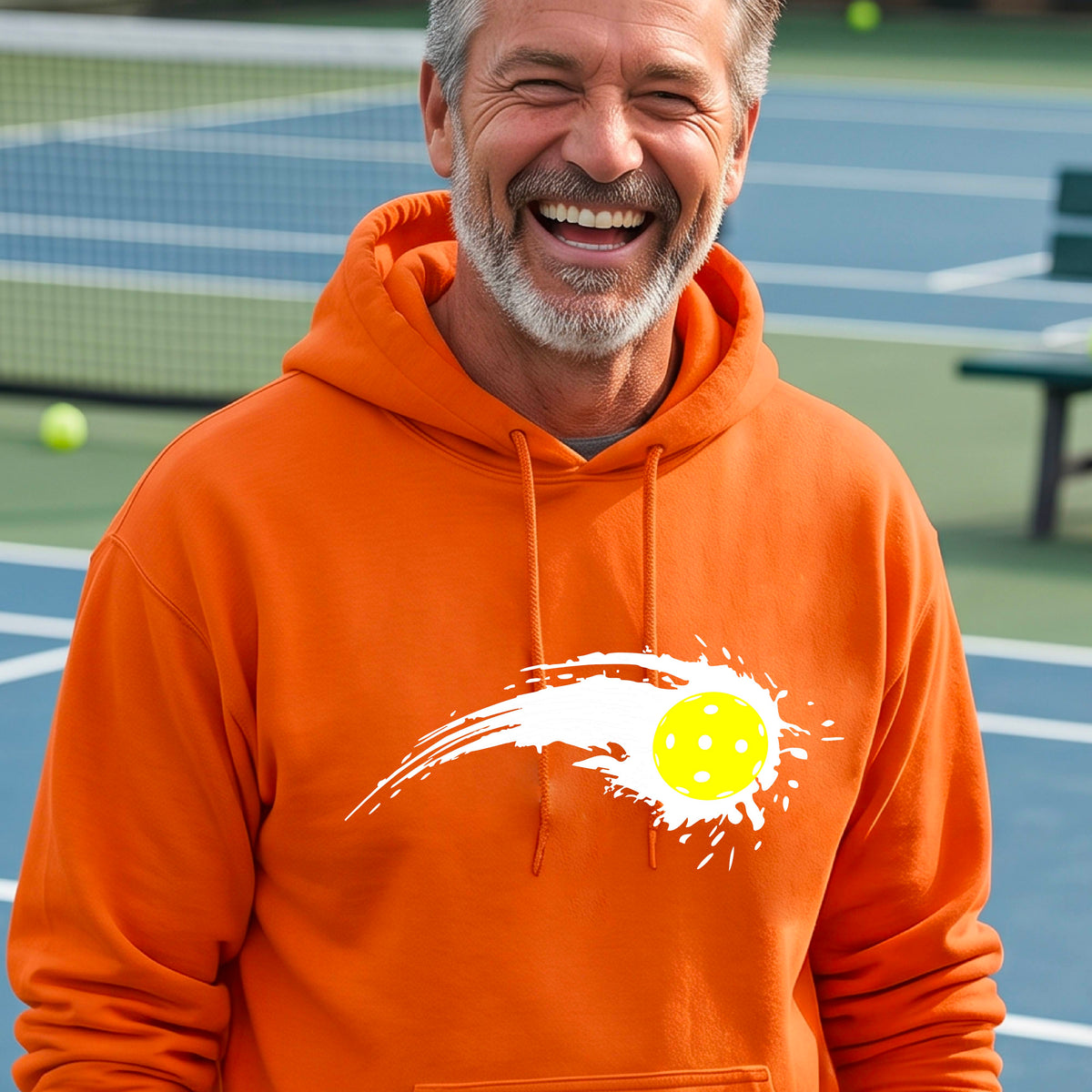 A man wearing an orange unisex hoodie pullover with a white pickleball graphic of a yellow pickleball with a motion trail effect, displayed in front of a pickleball court. 