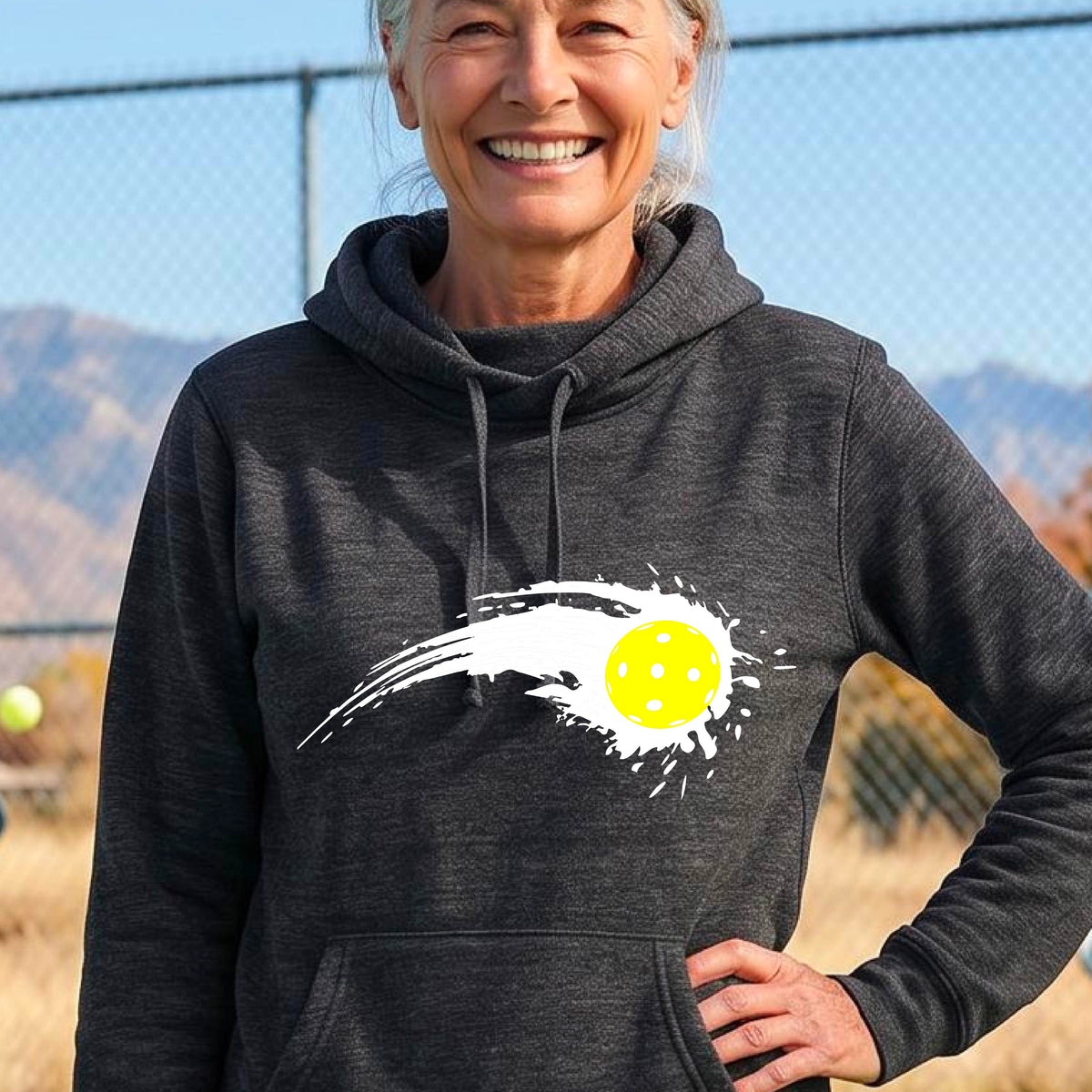 A woman wearing a black cowl-neck hoodie pullover with a white pickleball graphic of a yellow pickleball with a motion trail effect, displayed in front of a pickleball court. 