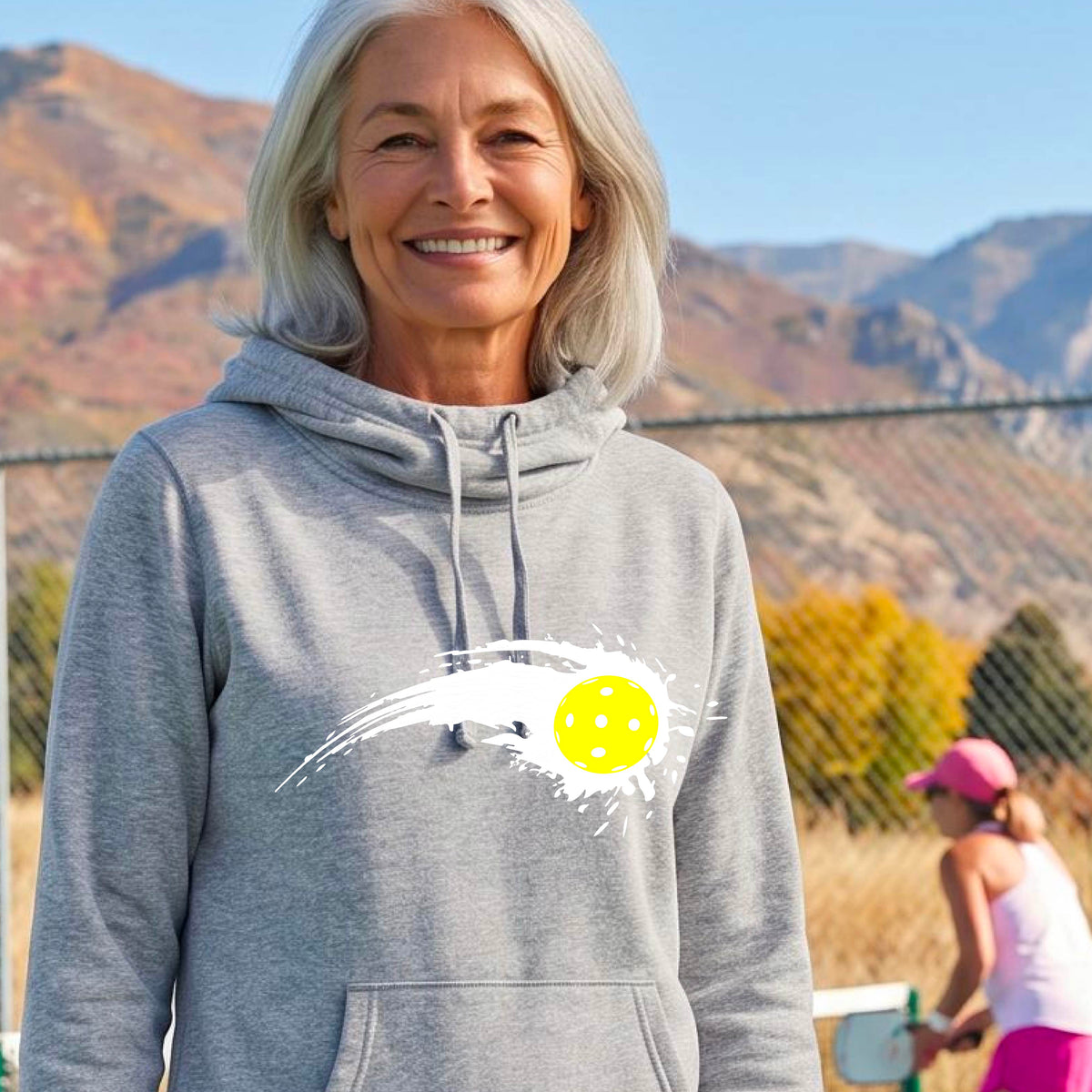 A woman wearing a light gray cowl-neck hoodie pullover with a white pickleball graphic of a yellow pickleball with a motion trail effect, displayed in front of a pickleball court. 