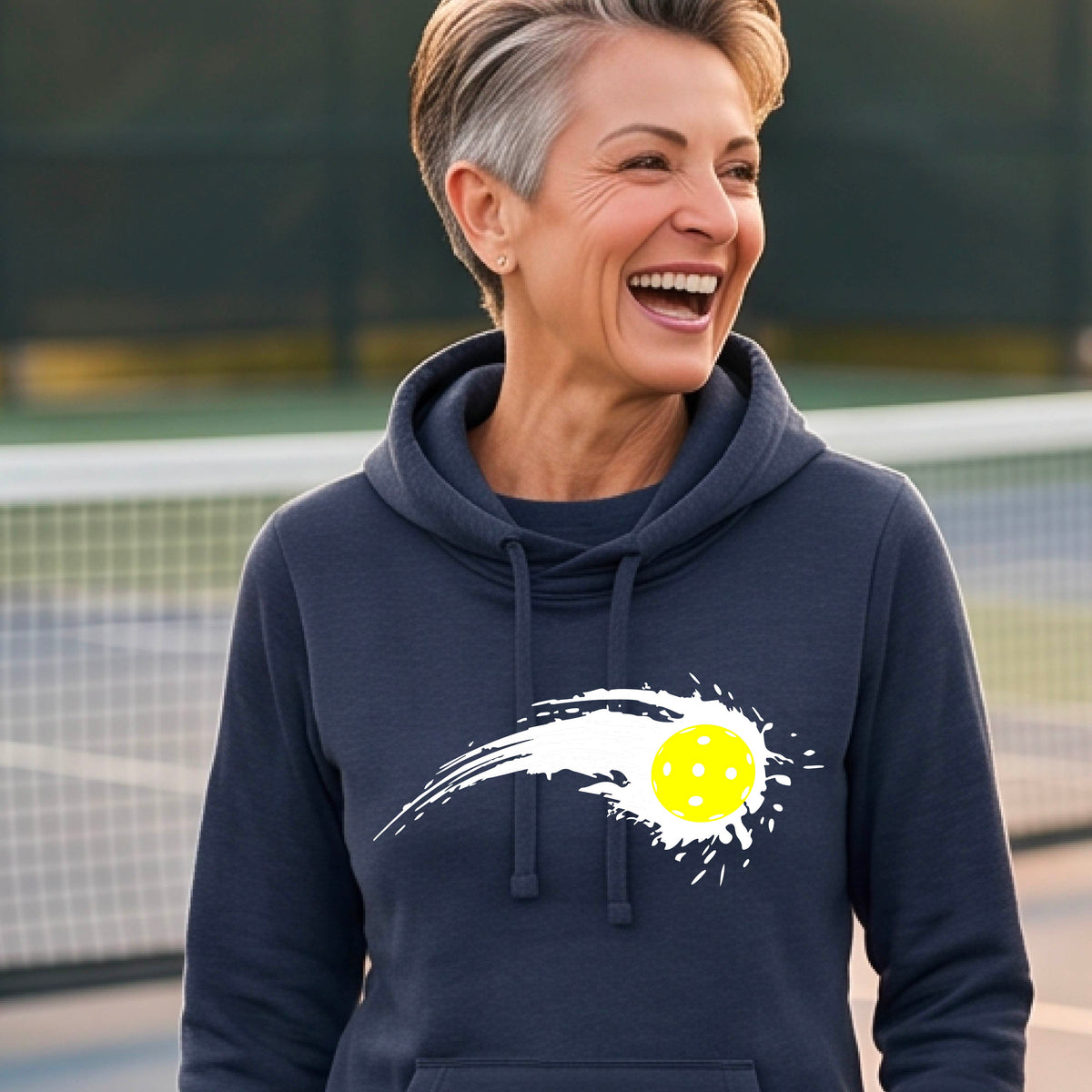 A woman wearing a navy blue cowl-neck hoodie pullover with a white pickleball graphic of a yellow pickleball with a motion trail effect, displayed in front of a pickleball court. 