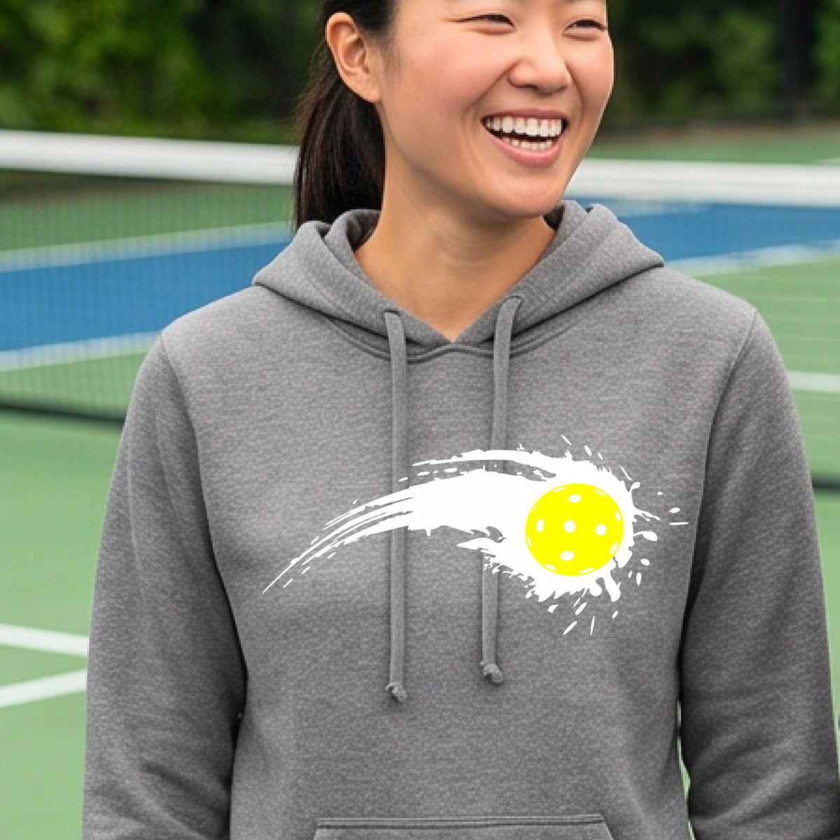 A woman wearing a gray fitted hoodie pullover with a white pickleball graphic of a yellow pickleball with a motion trail effect, displayed in front of a pickleball court. 