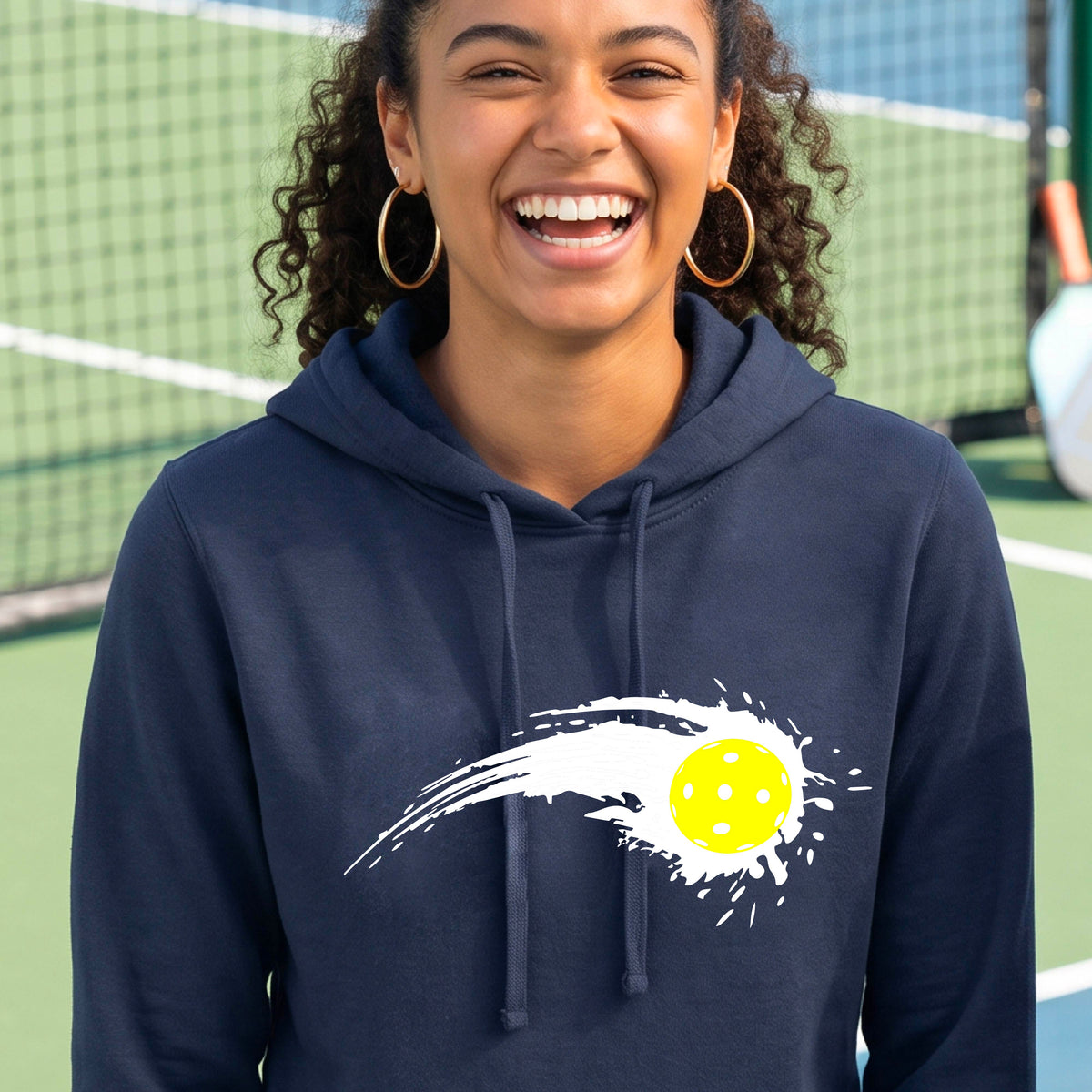 A woman wearing a navy blue fitted hoodie pullover with a white pickleball graphic of a yellow pickleball with a motion trail effect, displayed in front of a pickleball court. 