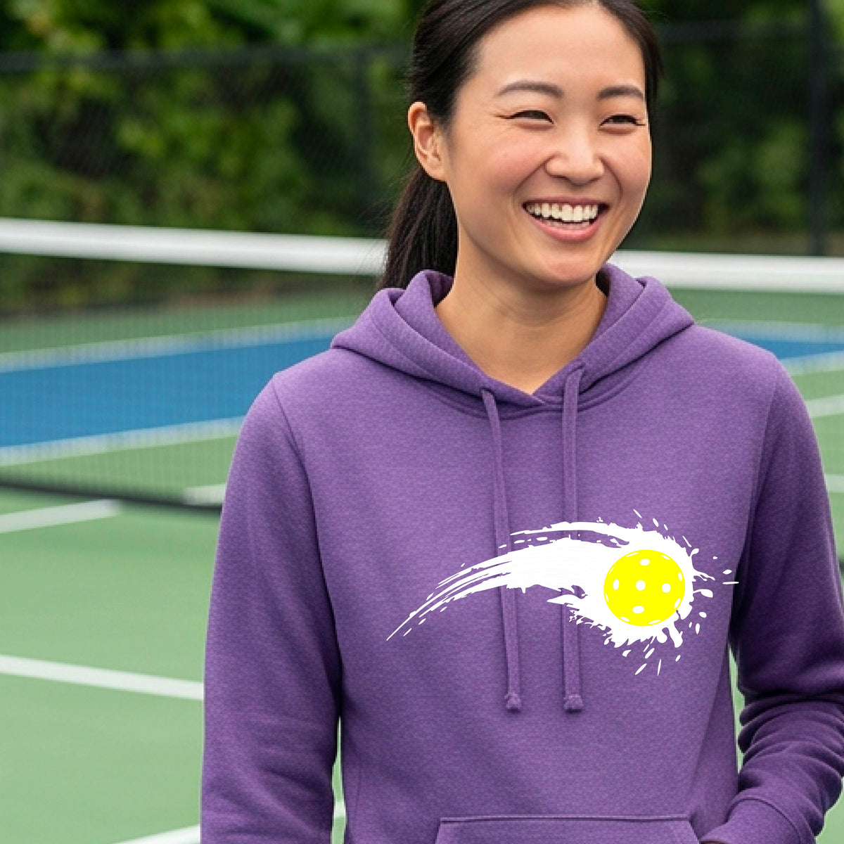A woman wearing a purple fitted hoodie pullover with a white pickleball graphic of a yellow pickleball with a motion trail effect, displayed in front of a pickleball court. 
