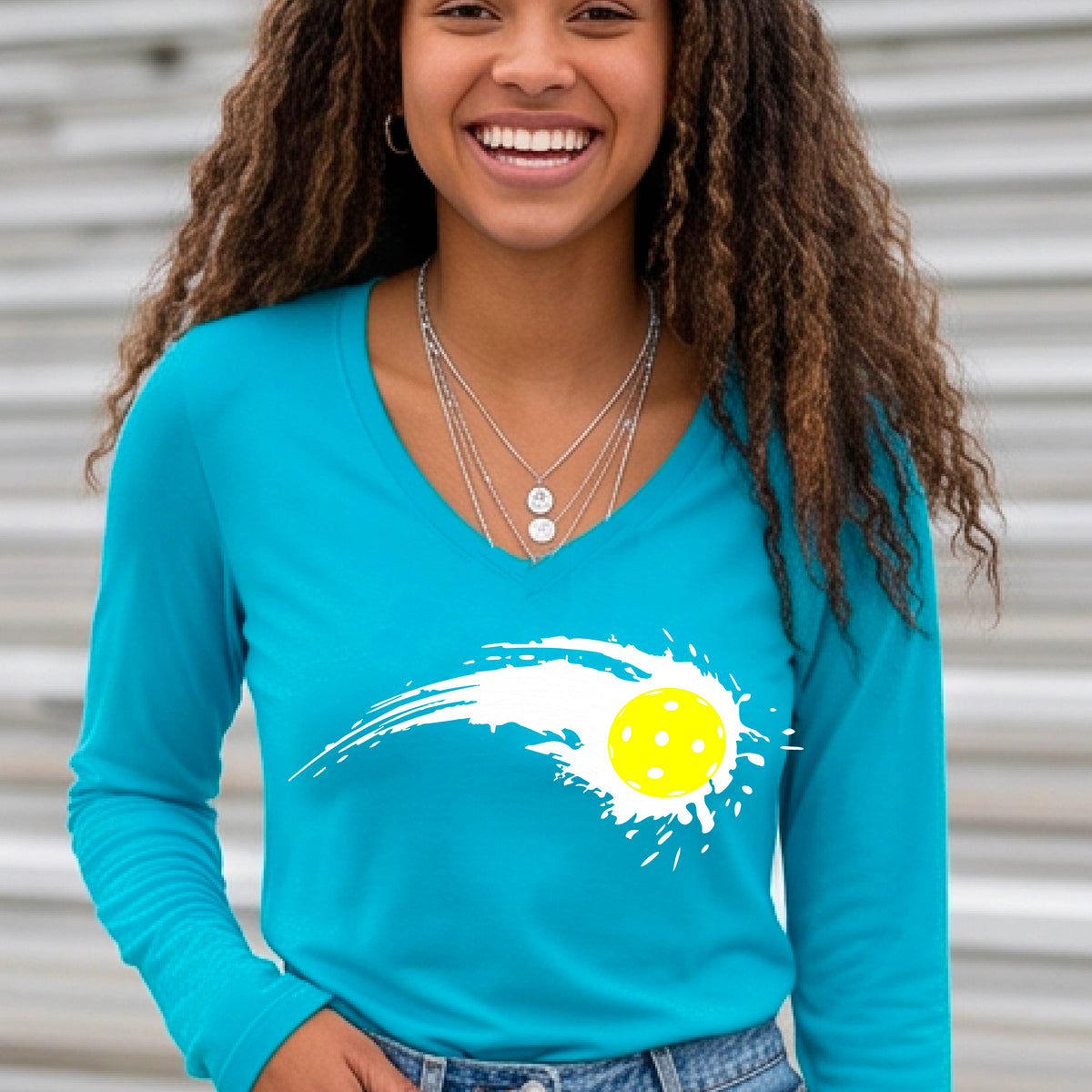 A woman wearing a bright blue long-sleeve T-shirt with a white pickleball graphic of a yellow pickleball with a motion trail effect, displayed in front of a pickleball court.