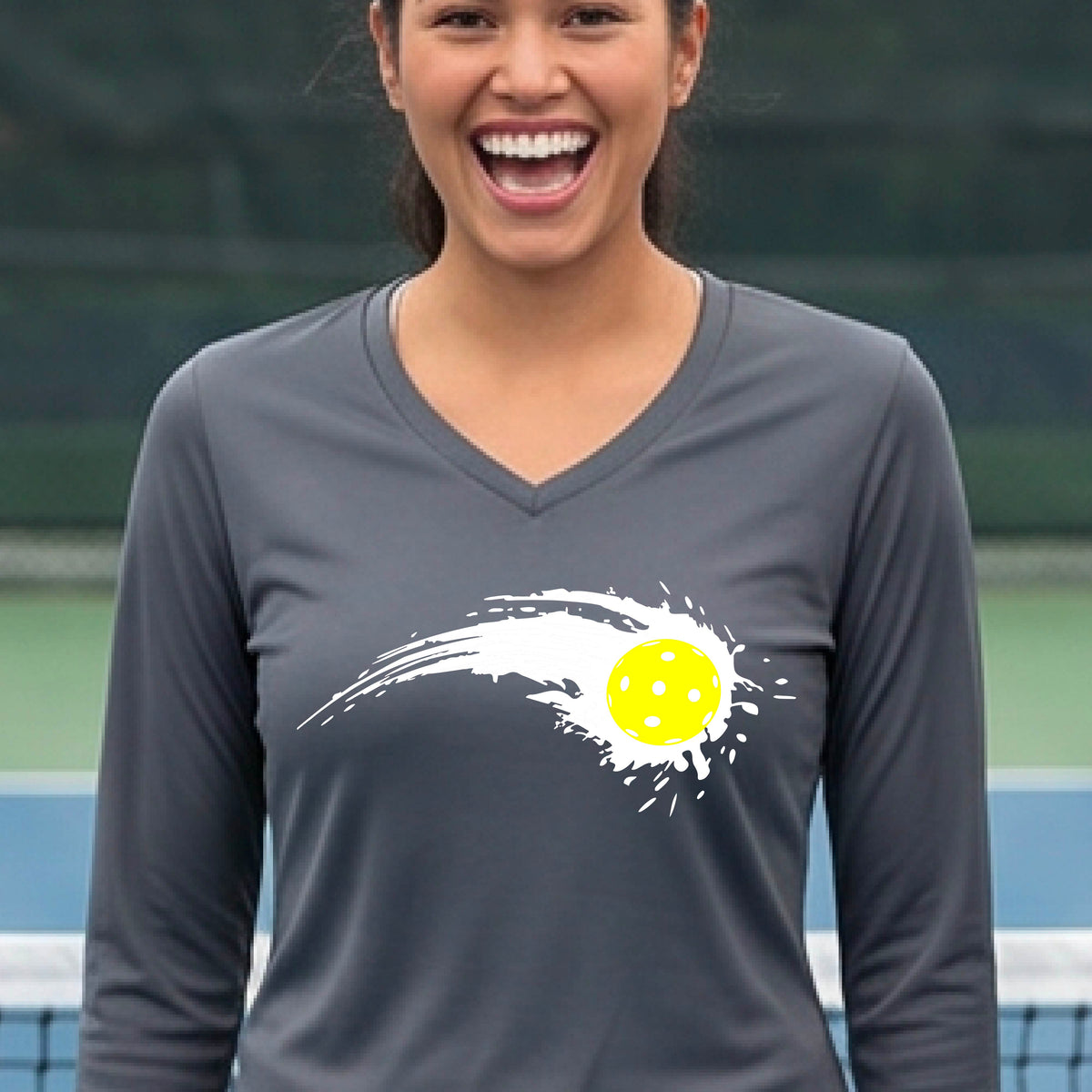 A woman wearing a dark gray long-sleeve T-shirt with a white pickleball graphic of a yellow pickleball with a motion trail effect, displayed in front of a pickleball court.