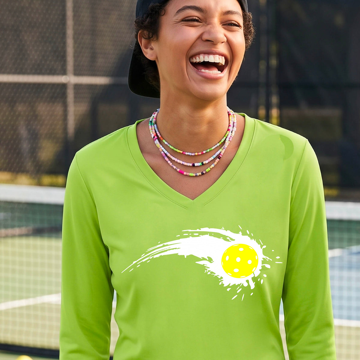 A woman wearing a lime green long-sleeve T-shirt with a white pickleball graphic of a yellow pickleball with a motion trail effect, displayed in front of a pickleball court.