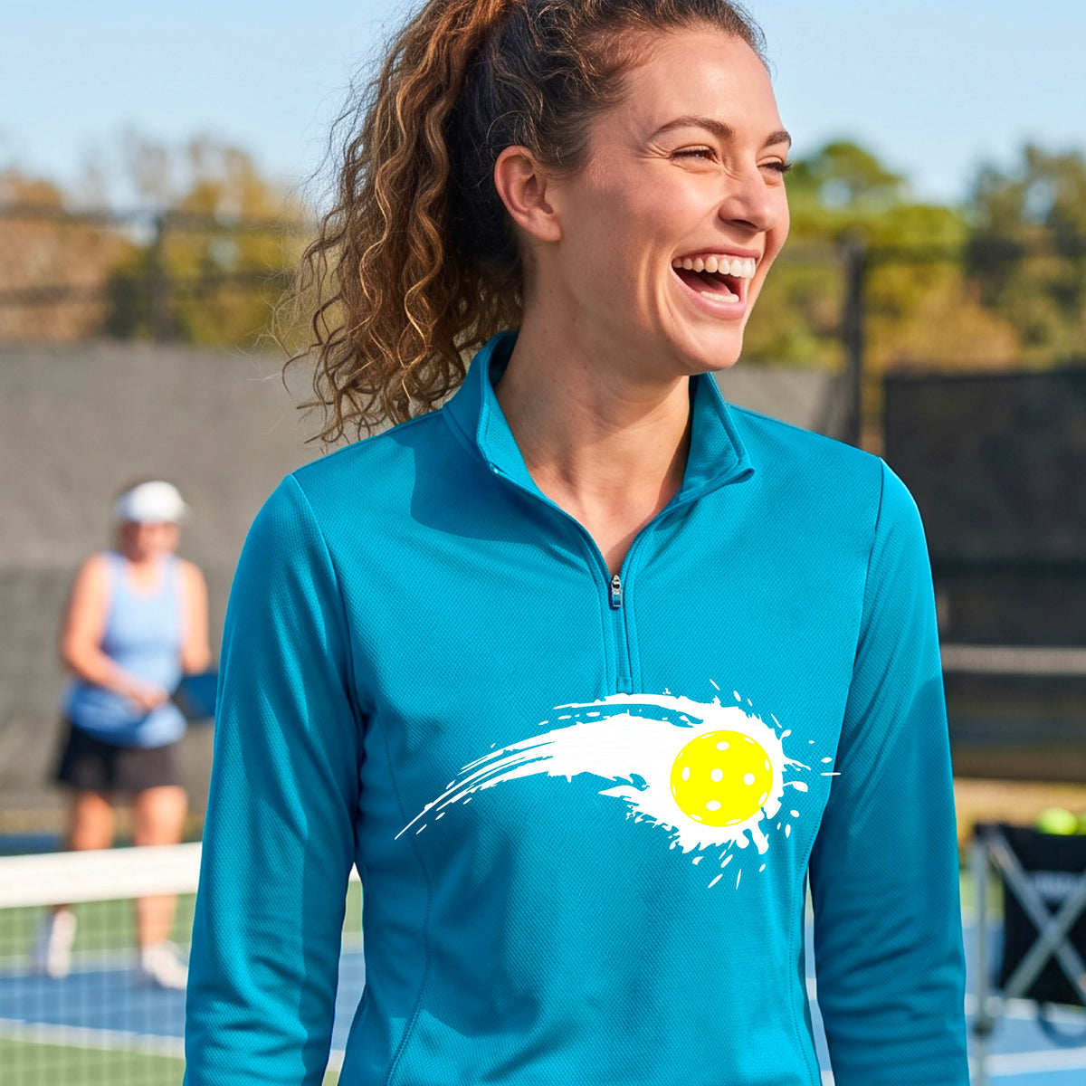 A close-up of a woman wearing a bright blue quarter-zip shirt with a white pickleball graphic of a yellow pickleball with a motion trail effect on the front.