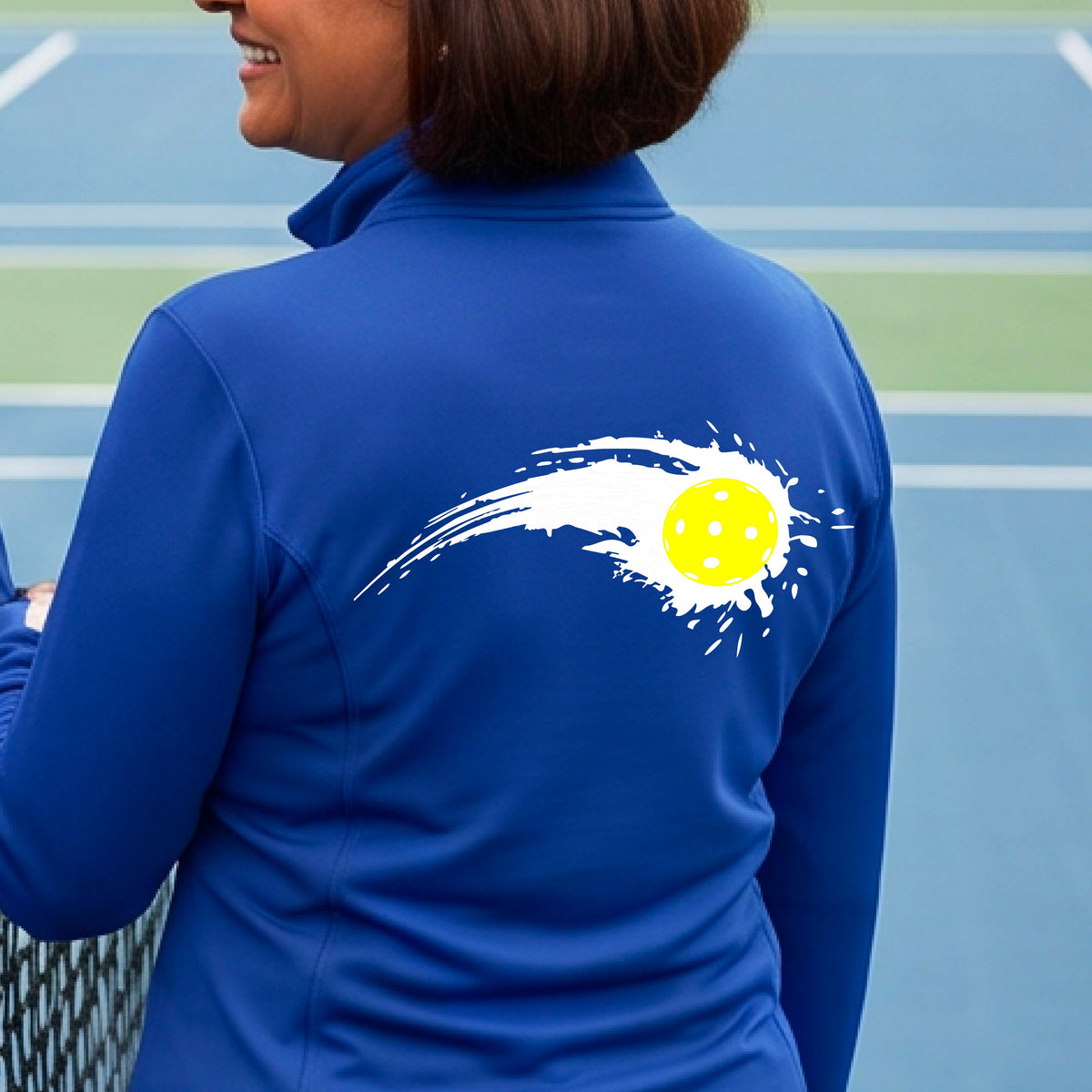 A close-up of a woman wearing a royal blue quarter-zip shirt with a white pickleball graphic of a yellow pickleball with a motion trail effect on the back