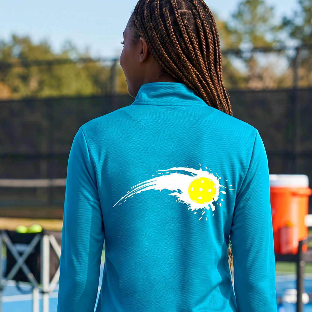 A close-up of a woman wearing a bright blue quarter-zip shirt with a white pickleball graphic of a yellow pickleball with a motion trail effect on the back.