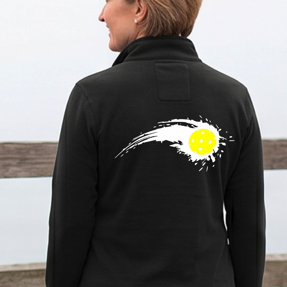 A close-up of a woman wearing a black quarter-zip shirt with a white pickleball graphic of a yellow pickleball with a motion trail effect on the back.