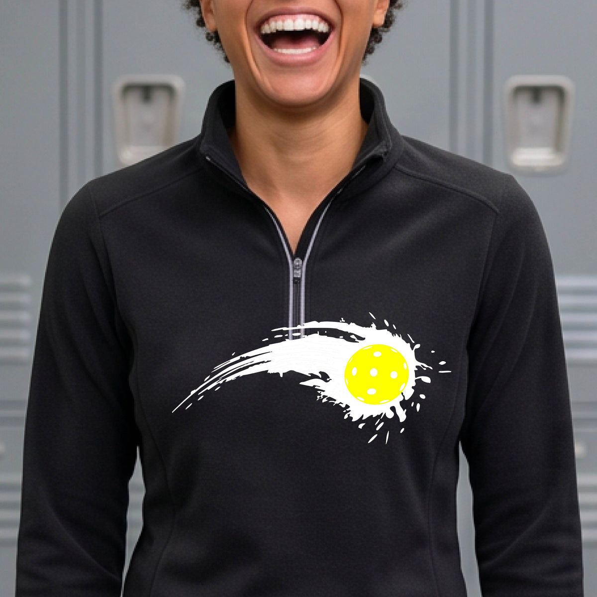 A close-up of a woman wearing a black quarter-zip shirt with a white pickleball graphic of a yellow pickleball with a motion trail effect on the front,.