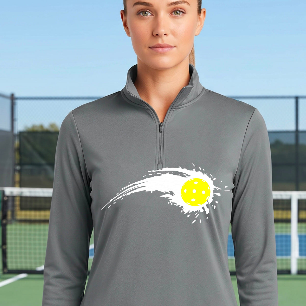 A close-up of a woman wearing a gray quarter-zip shirt with a white pickleball graphic of a yellow pickleball with a motion trail effect on the front.