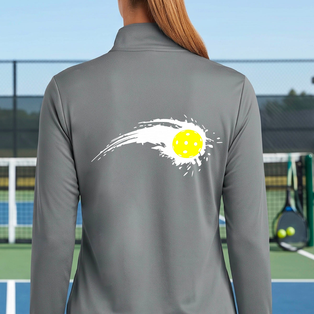 A close-up of a woman wearing a gray quarter-zip shirt with a white pickleball graphic of a yellow pickleball with a motion trail effect on the back.