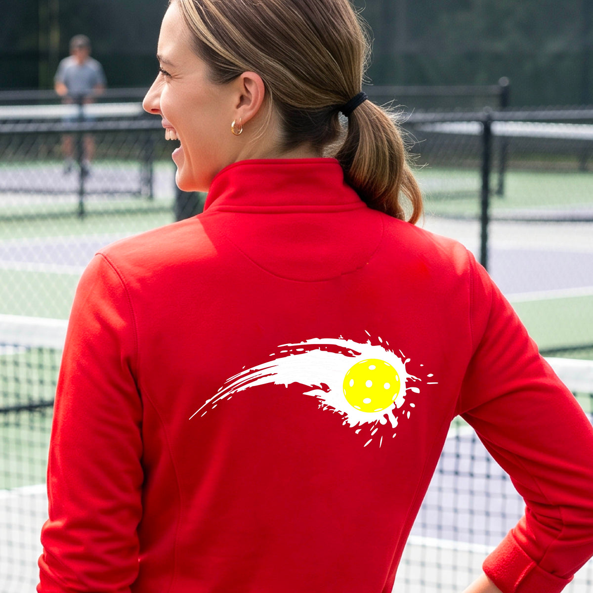 A close-up of a woman wearing a red quarter-zip shirt with a white pickleball graphic of a yellow pickleball with a motion trail effect on the back.