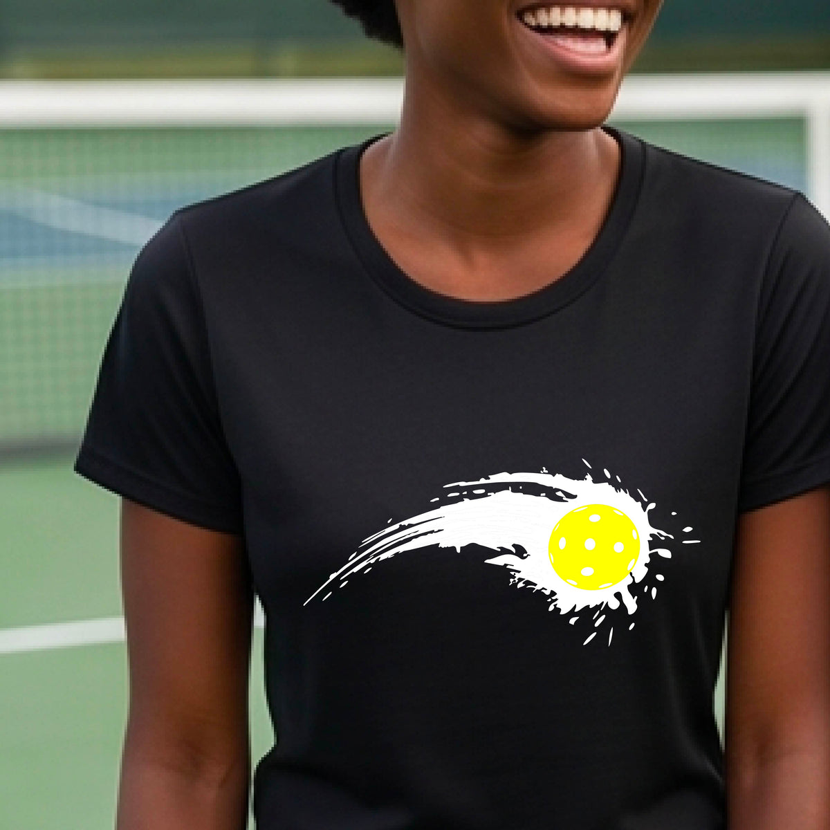 A woman wearing a black short-sleeve T-shirt with a white pickleball graphic of a yellow pickleball with a motion trail effect, displayed in front of a pickleball court. 