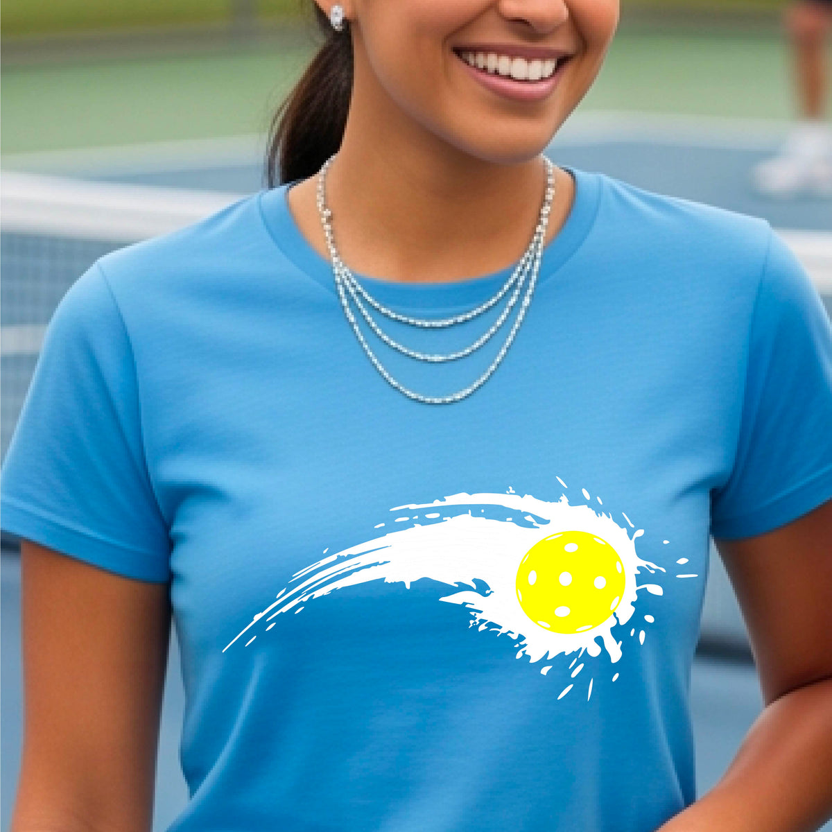 A woman wearing a light blue short-sleeve T-shirt with a white pickleball graphic of a yellow pickleball with a motion trail effect, displayed in front of a pickleball court. 