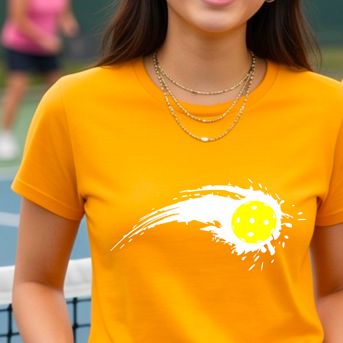 A woman wearing a mustard yellow gold short-sleeve T-shirt with a white pickleball graphic of a yellow pickleball with a motion trail effect, displayed in front of a pickleball court. 