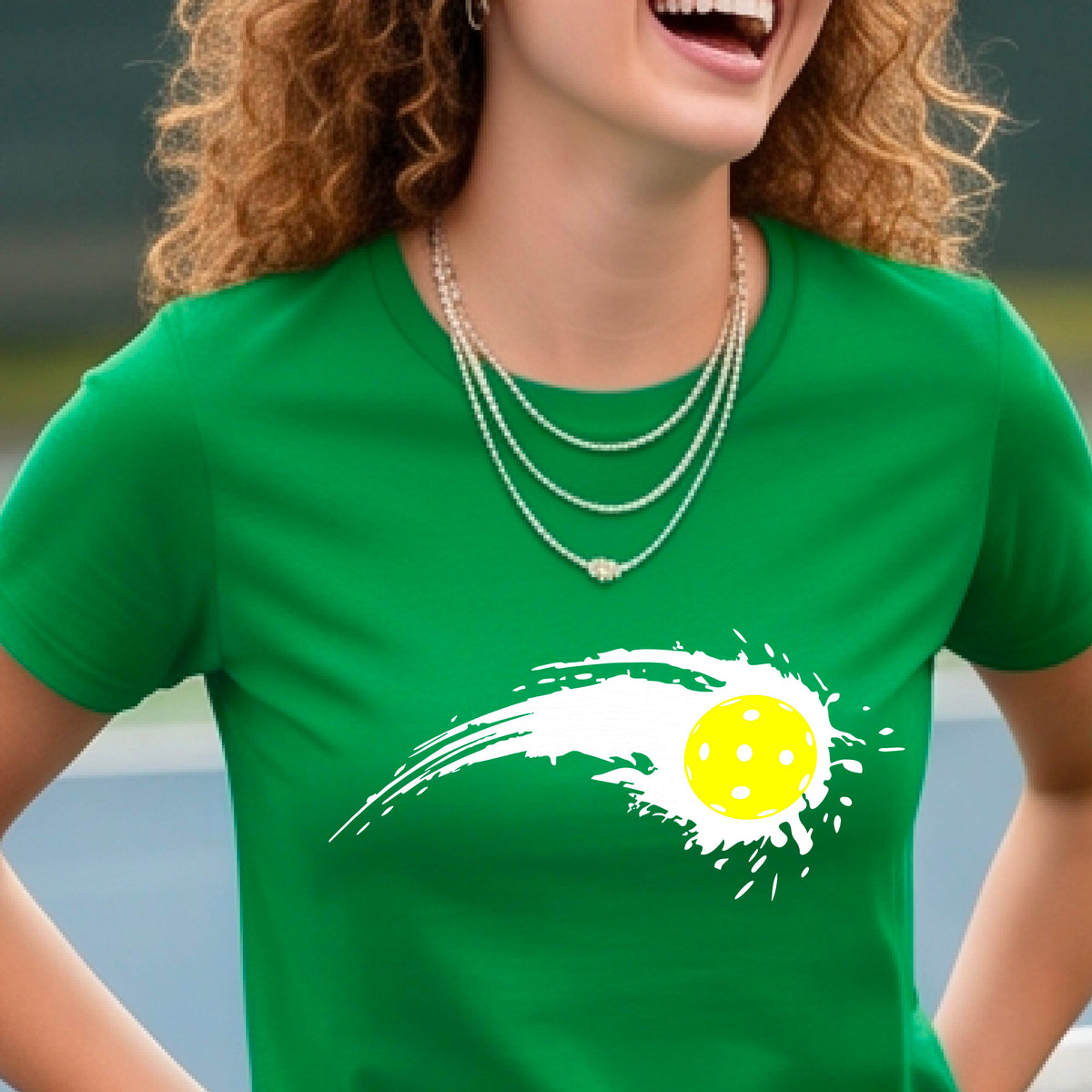 A woman wearing a green short-sleeve T-shirt with a white pickleball graphic of a yellow pickleball with a motion trail effect, displayed in front of a pickleball court. 