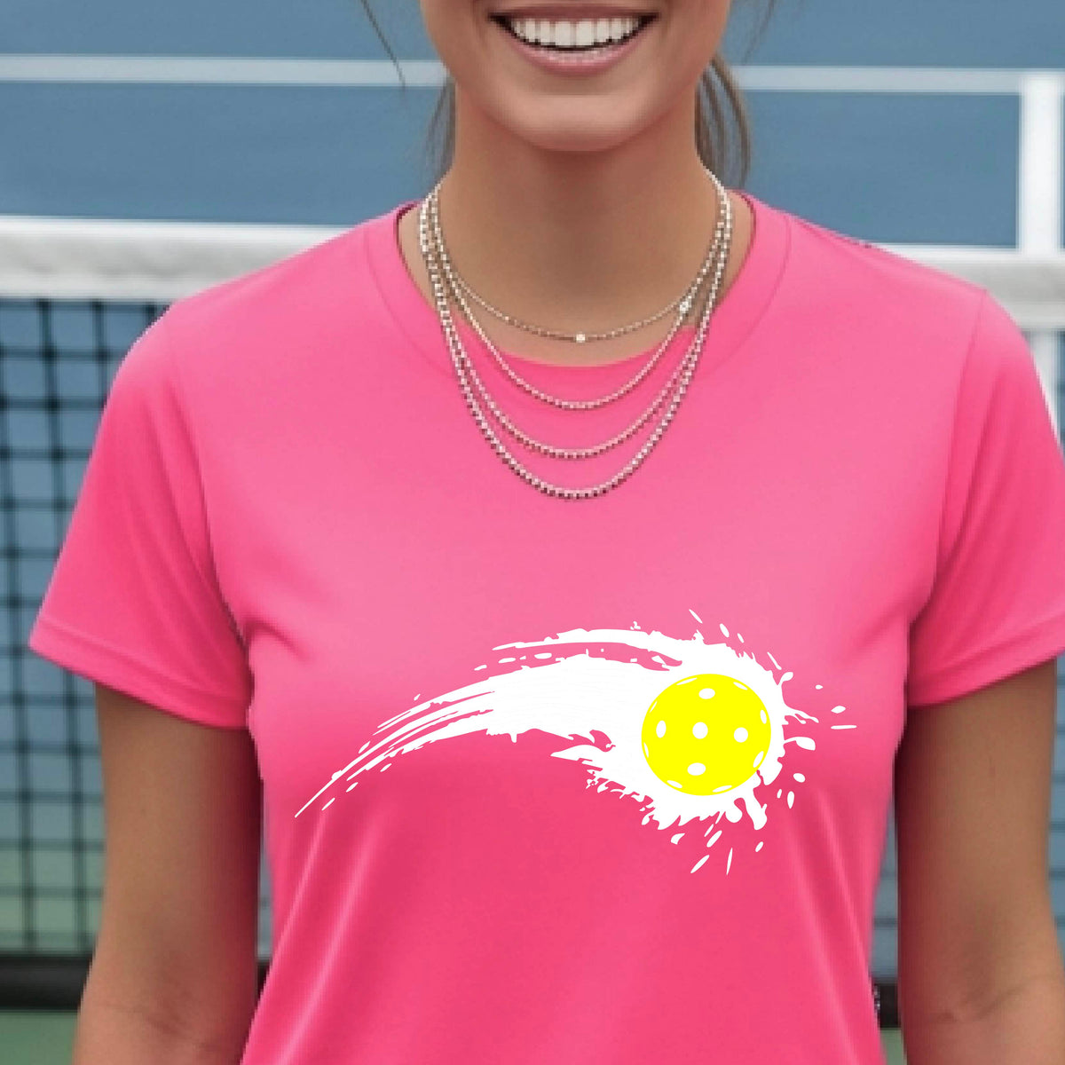 A woman wearing a bright pink short-sleeve T-shirt with a white pickleball graphic of a yellow pickleball with a motion trail effect, displayed in front of a pickleball court. 