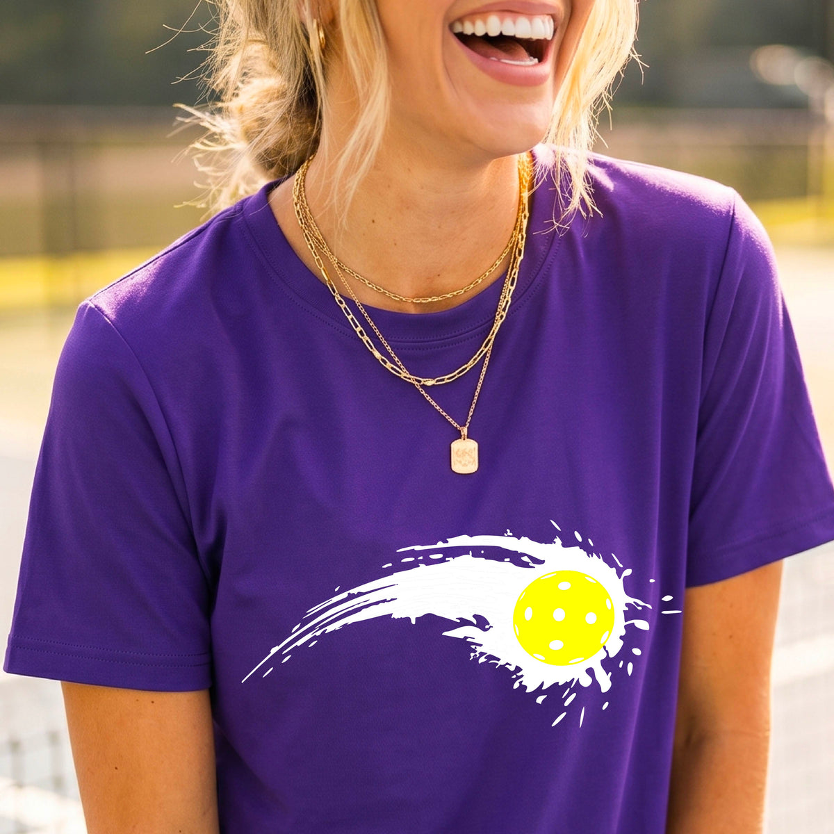 A woman wearing a purple short-sleeve T-shirt with a white pickleball graphic of a yellow pickleball with a motion trail effect, displayed in front of a pickleball court. 