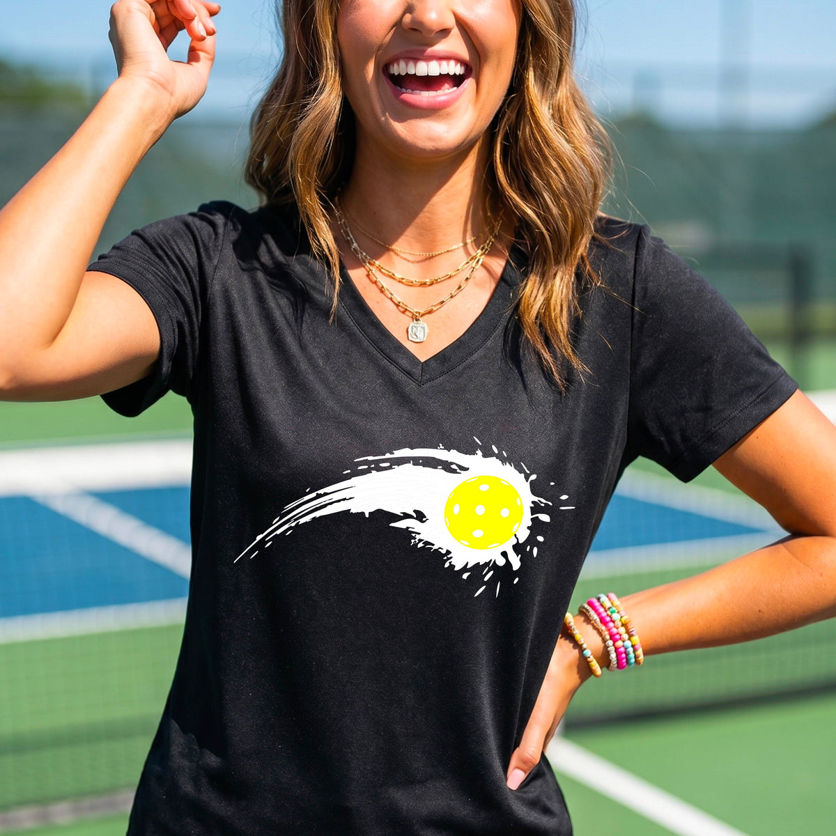 A woman wearing a black short-sleeve T-shirt with a white pickleball graphic of a yellow pickleball with a motion trail effect, displayed in front of a pickleball court. 