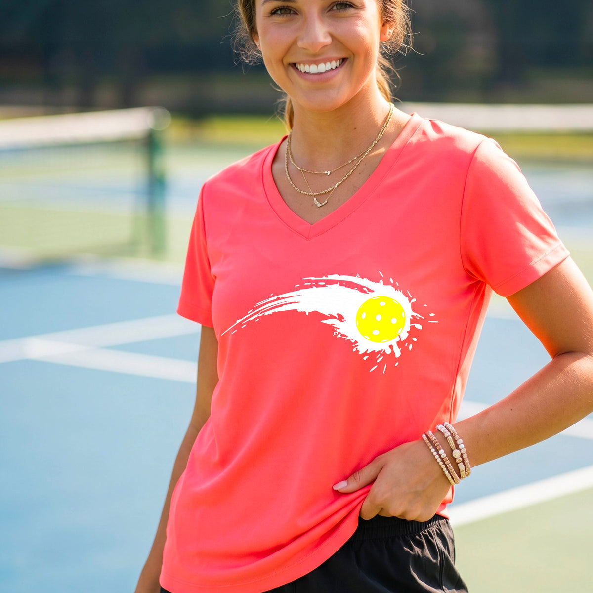 A woman wearing a coral pink short-sleeve T-shirt with a white pickleball graphic of a yellow pickleball with a motion trail effect, displayed in front of a pickleball court. 