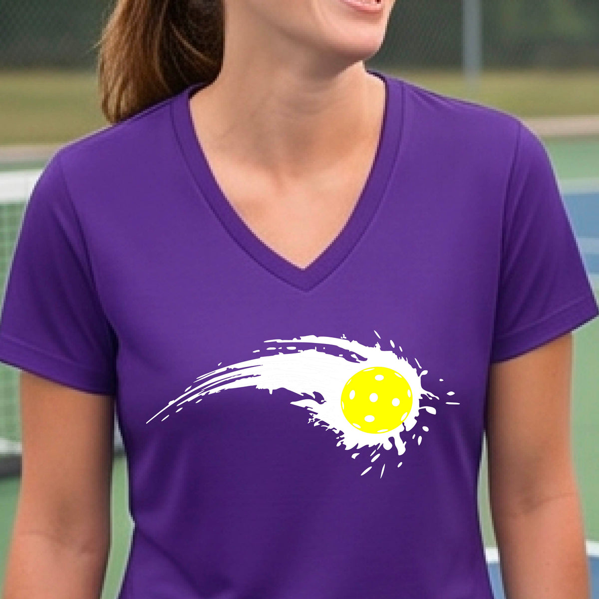 A woman wearing a purple short-sleeve T-shirt with a white pickleball graphic of a yellow pickleball with a motion trail effect, displayed in front of a pickleball court. 