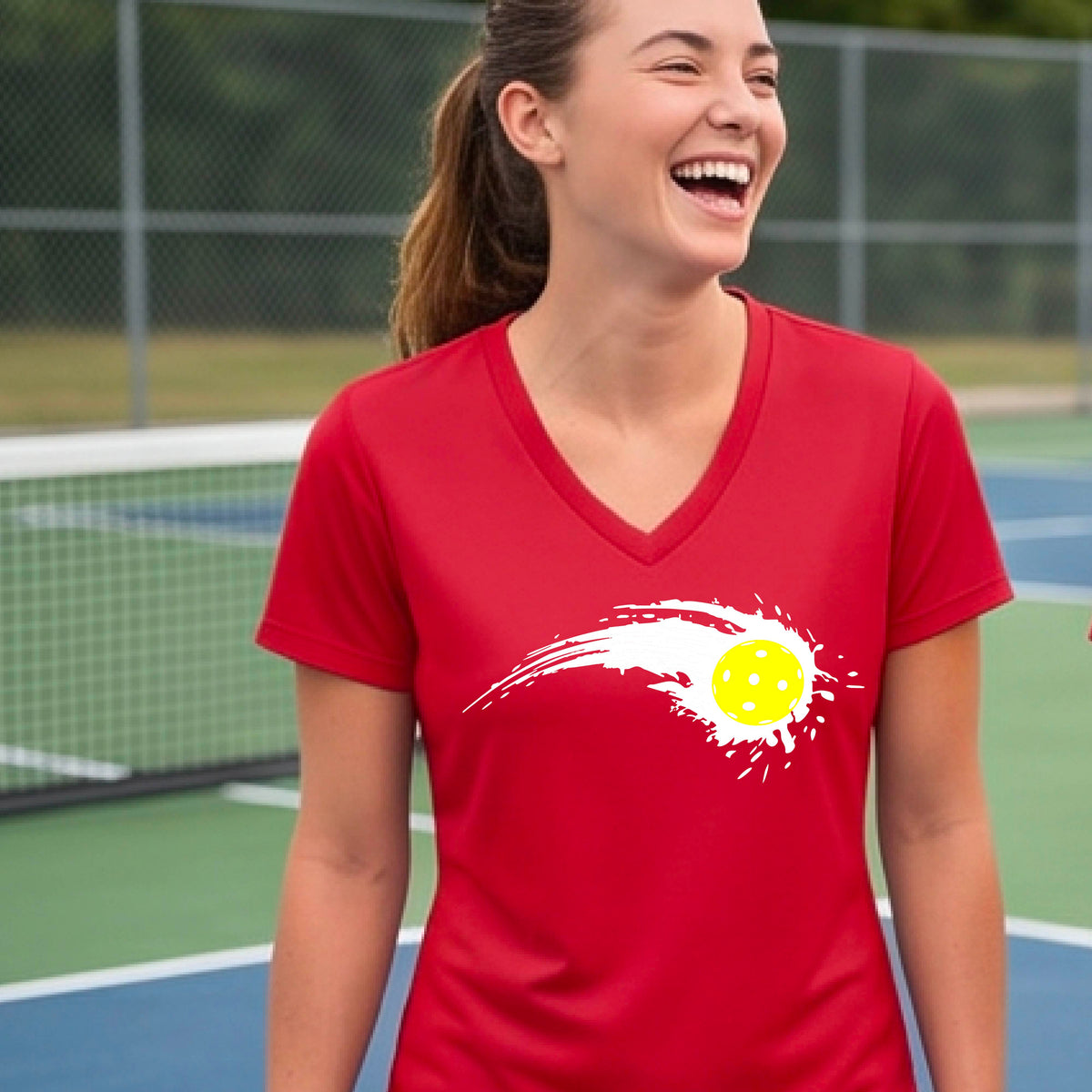 A woman wearing a red short-sleeve T-shirt with a white pickleball graphic of a yellow pickleball with a motion trail effect, displayed in front of a pickleball court. 