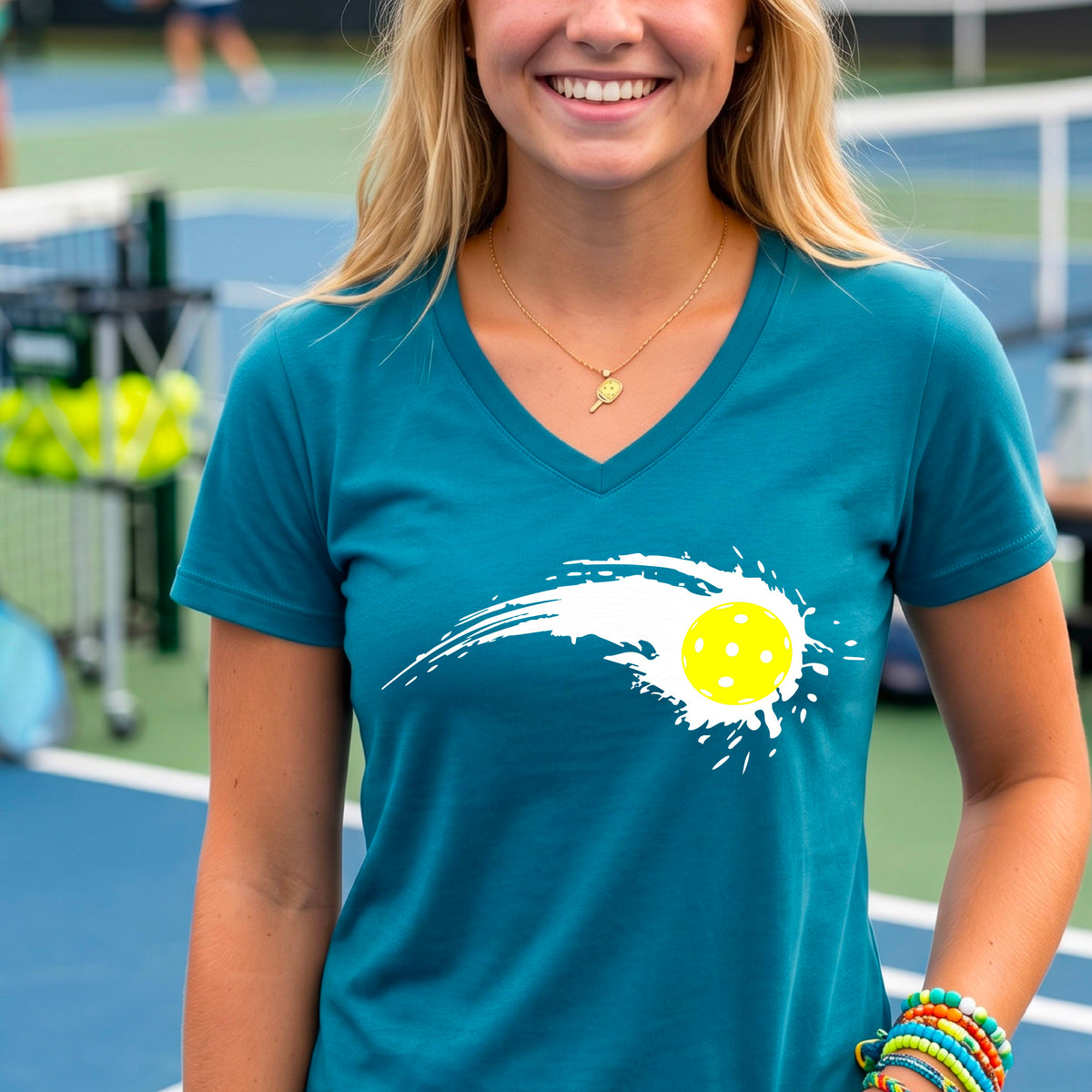 A woman wearing a teal short-sleeve T-shirt with a white pickleball graphic of a yellow pickleball with a motion trail effect, displayed in front of a pickleball court. 