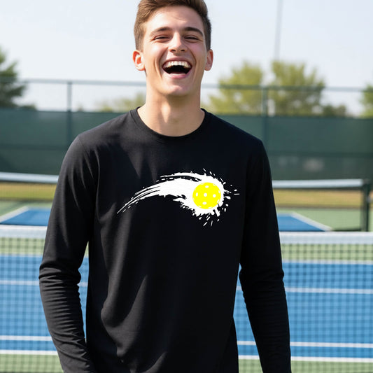 A close-up of a young person wearing a black long-sleeve t-shirt with a white pickleball graphic of a yellow pickleball with a motion trail effect, displayed in front of a pickleball court. 