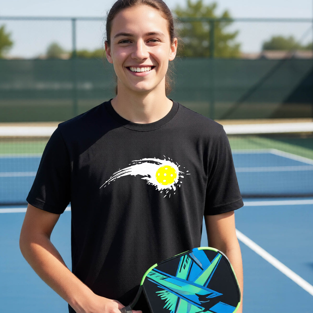A teenager wearing a black short-sleeve T-shirt with a white pickleball graphic of a yellow pickleball with a motion trail effect, displayed in front of a pickleball court. 