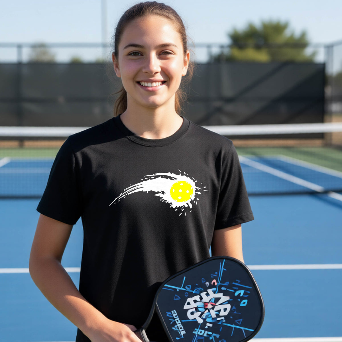 A teenager wearing a black short-sleeve T-shirt with a white pickleball graphic of a yellow pickleball with a motion trail effect, displayed in front of a pickleball court. 