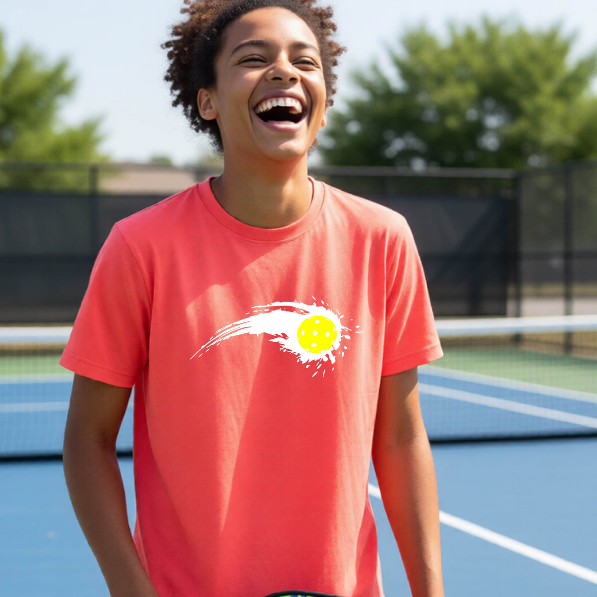 A teenager wearing a coral pink short-sleeve T-shirt with a white pickleball graphic of a yellow pickleball with a motion trail effect, displayed in front of a pickleball court. 