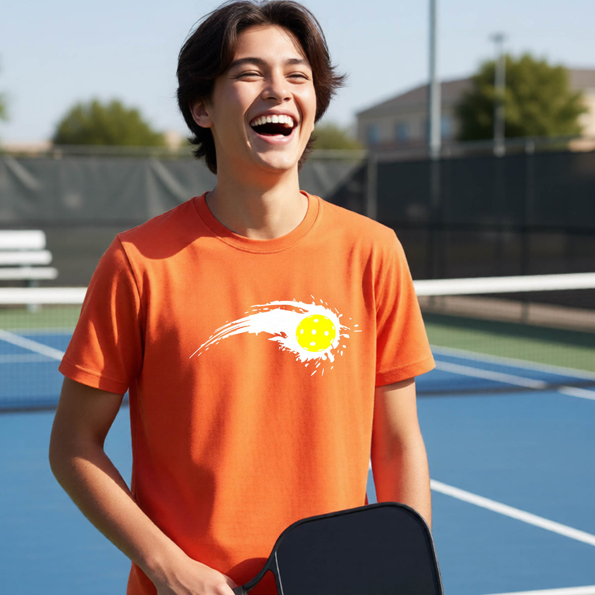 A teenager wearing an orange short-sleeve T-shirt with a white pickleball graphic of a yellow pickleball with a motion trail effect, displayed in front of a pickleball court. 