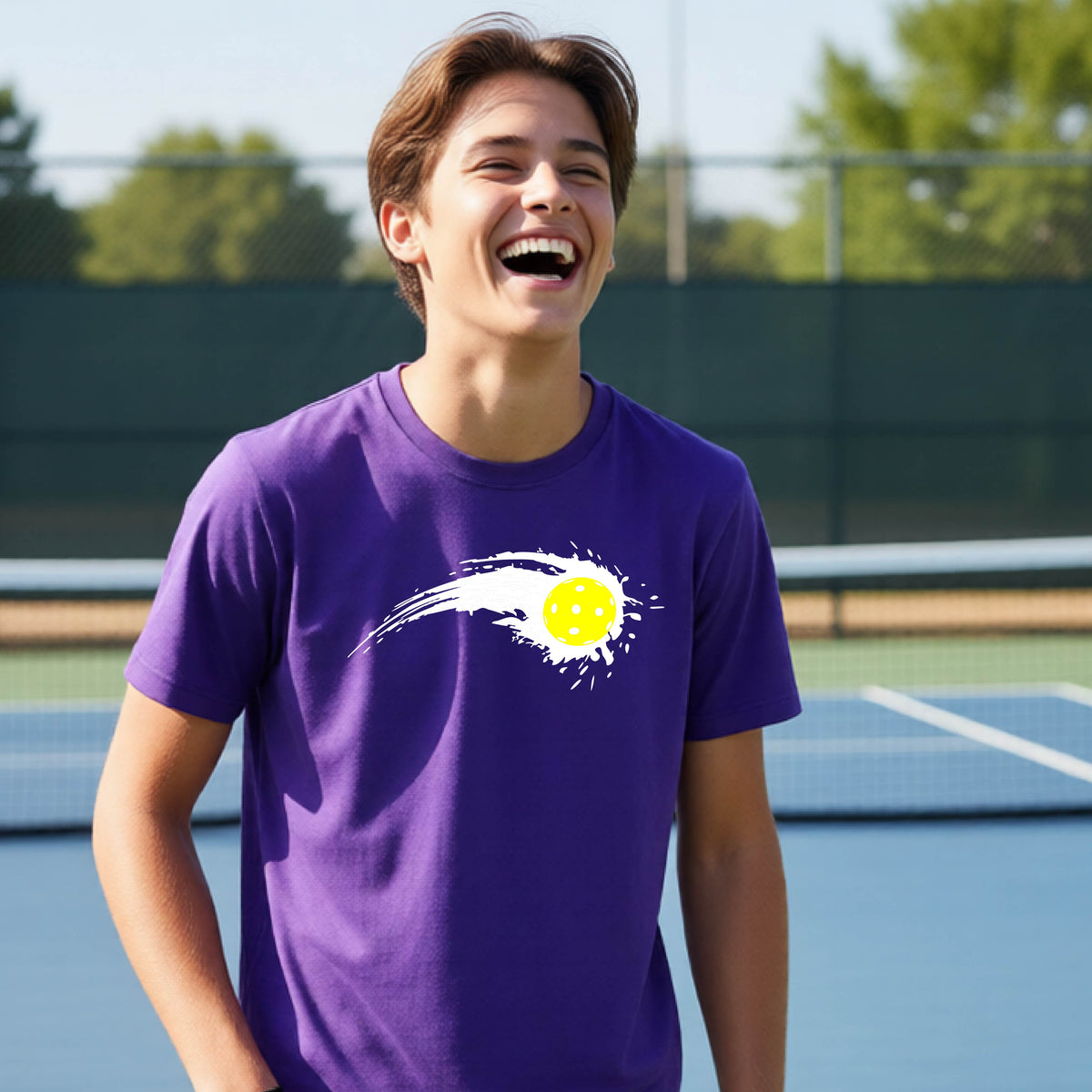 A teenager wearing a purple short-sleeve T-shirt with a white pickleball graphic of a yellow pickleball with a motion trail effect, displayed in front of a pickleball court. 