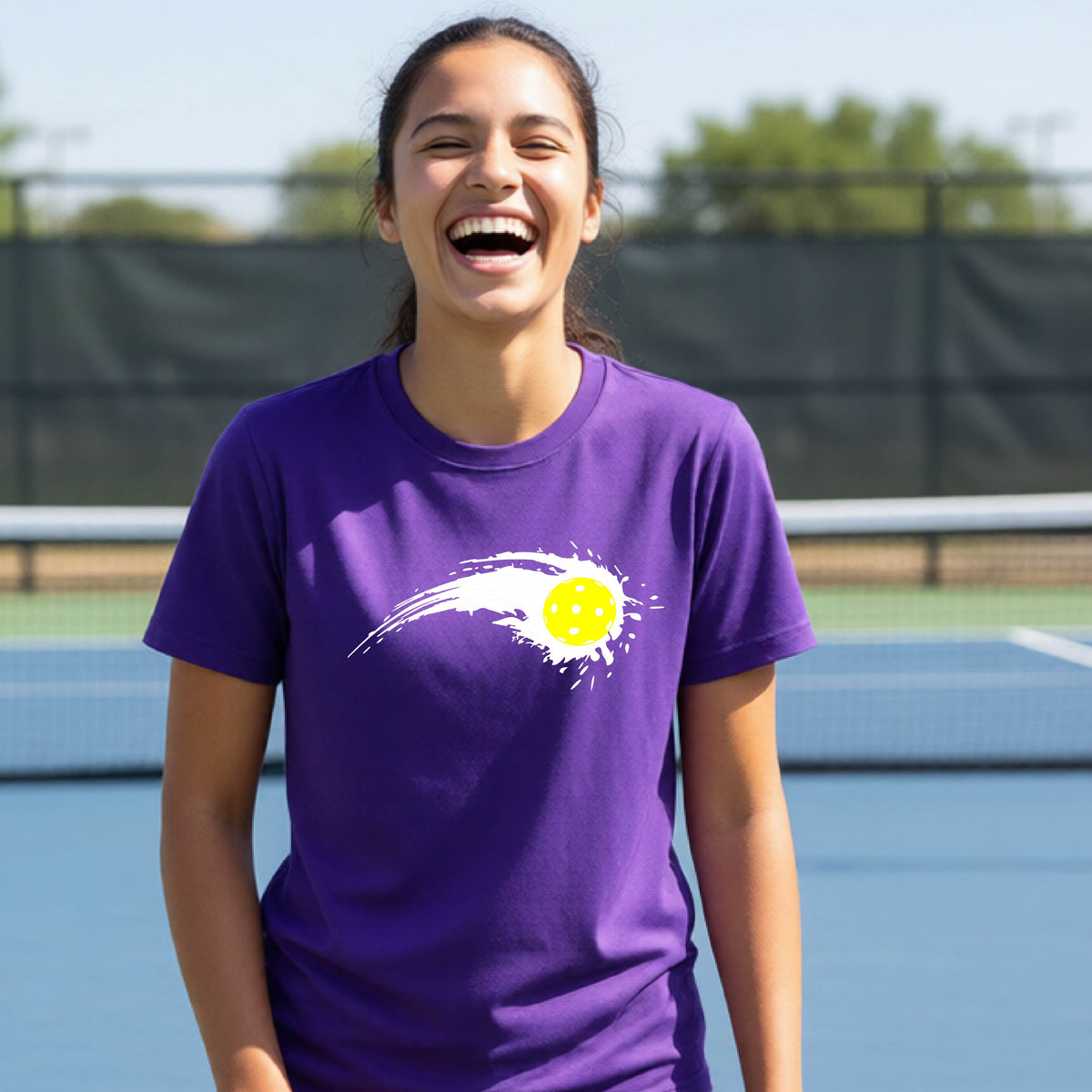 A teenager wearing a purple short-sleeve T-shirt with a white pickleball graphic of a yellow pickleball with a motion trail effect, displayed in front of a pickleball court. 