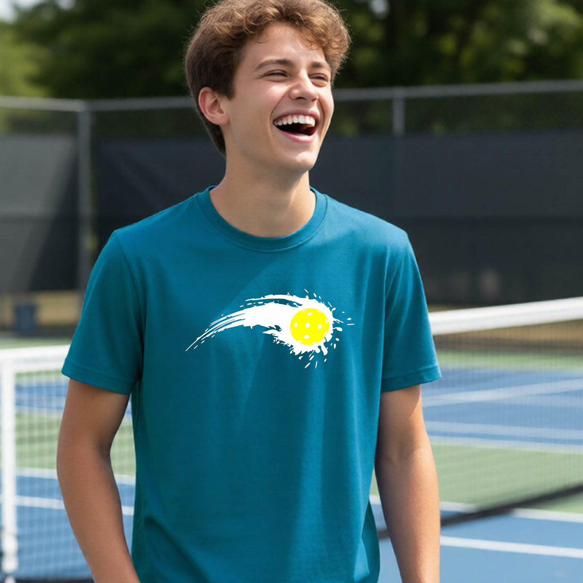 A teenager wearing a black short-sleeve T-shirt with a white pickleball graphic of a yellow pickleball with a motion trail effect, displayed in front of a pickleball court. 