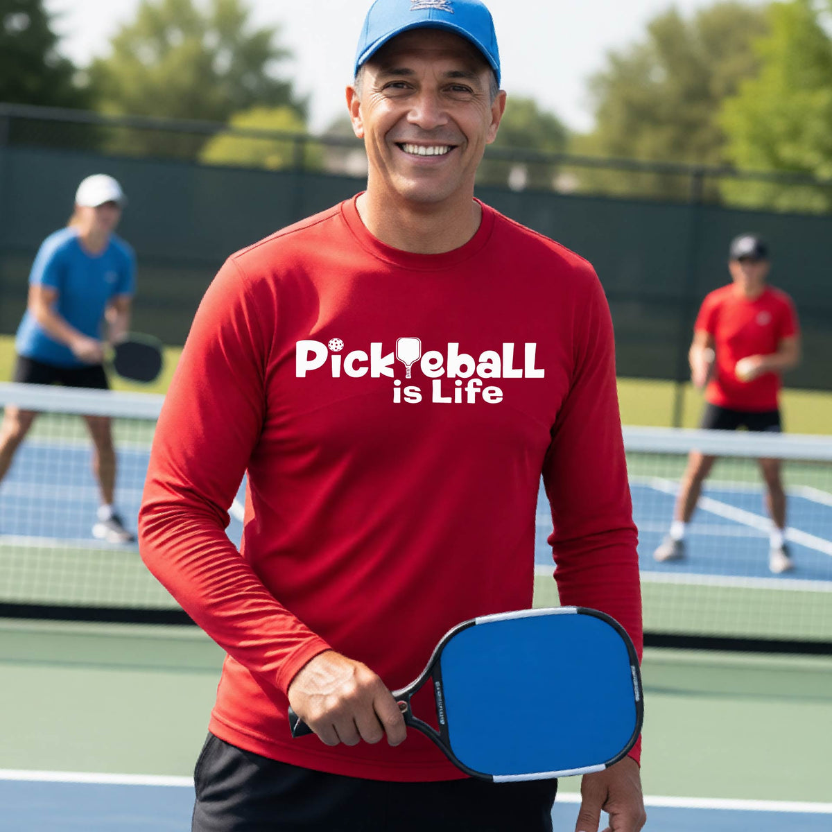 A smiling man on a pickleball court wearing a red long-sleeve shirt with the text 'Pickleball is Life' .
