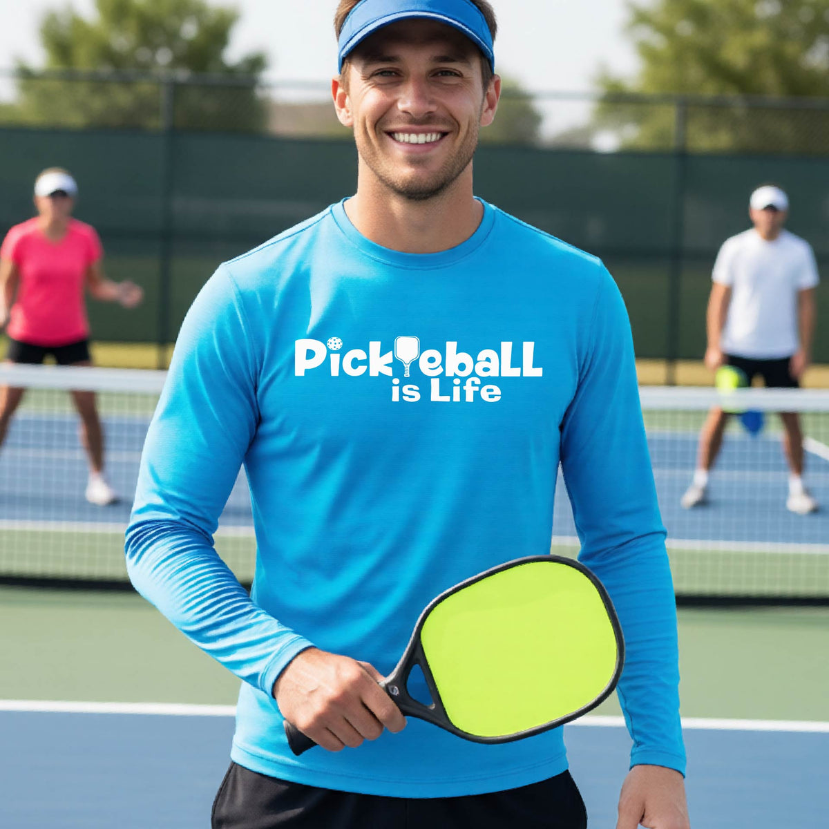 A smiling man on a pickleball court wearing a bright blue long-sleeve shirt with the text 'Pickleball is Life' .