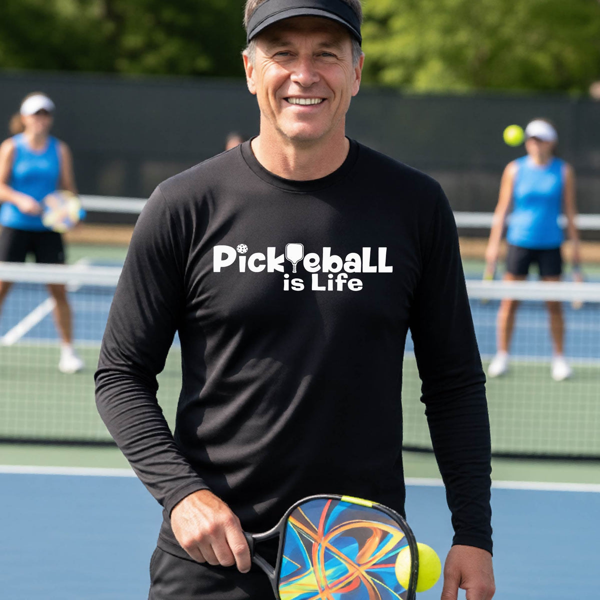 A smiling man on a pickleball court wearing a black long-sleeve shirt with the text 'Pickleball is Life' .