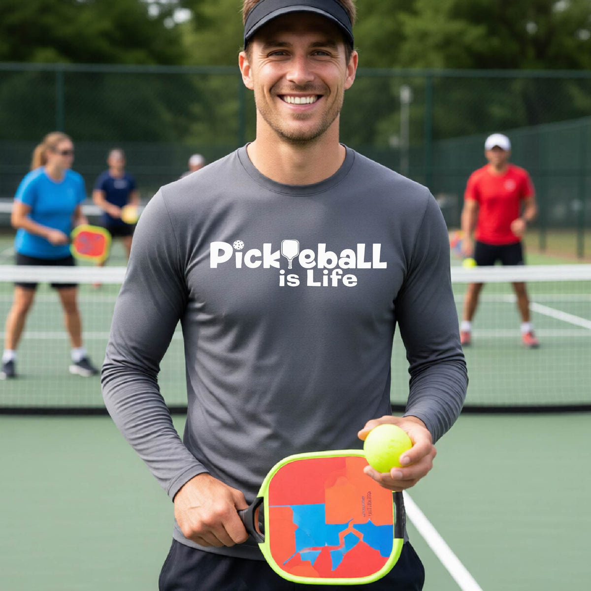 A smiling man on a pickleball court wearing a gray long-sleeve shirt with the text 'Pickleball is Life' .