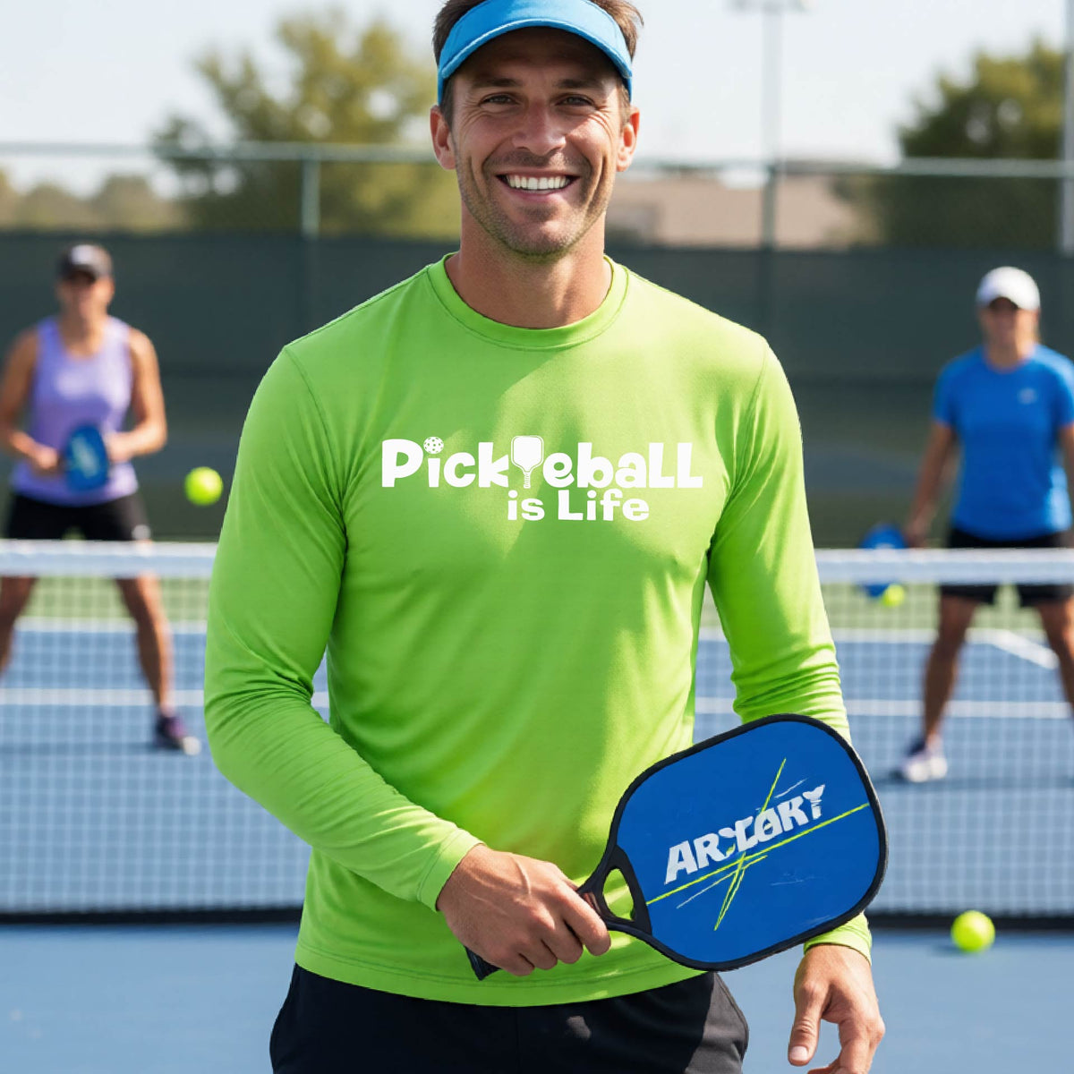 A smiling man on a pickleball court wearing a lime green long-sleeve shirt with the text 'Pickleball is Life' .