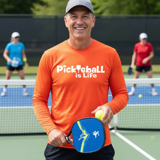 A smiling man on a pickleball court wearing an orange long-sleeve shirt with the text 'Pickleball is Life' .